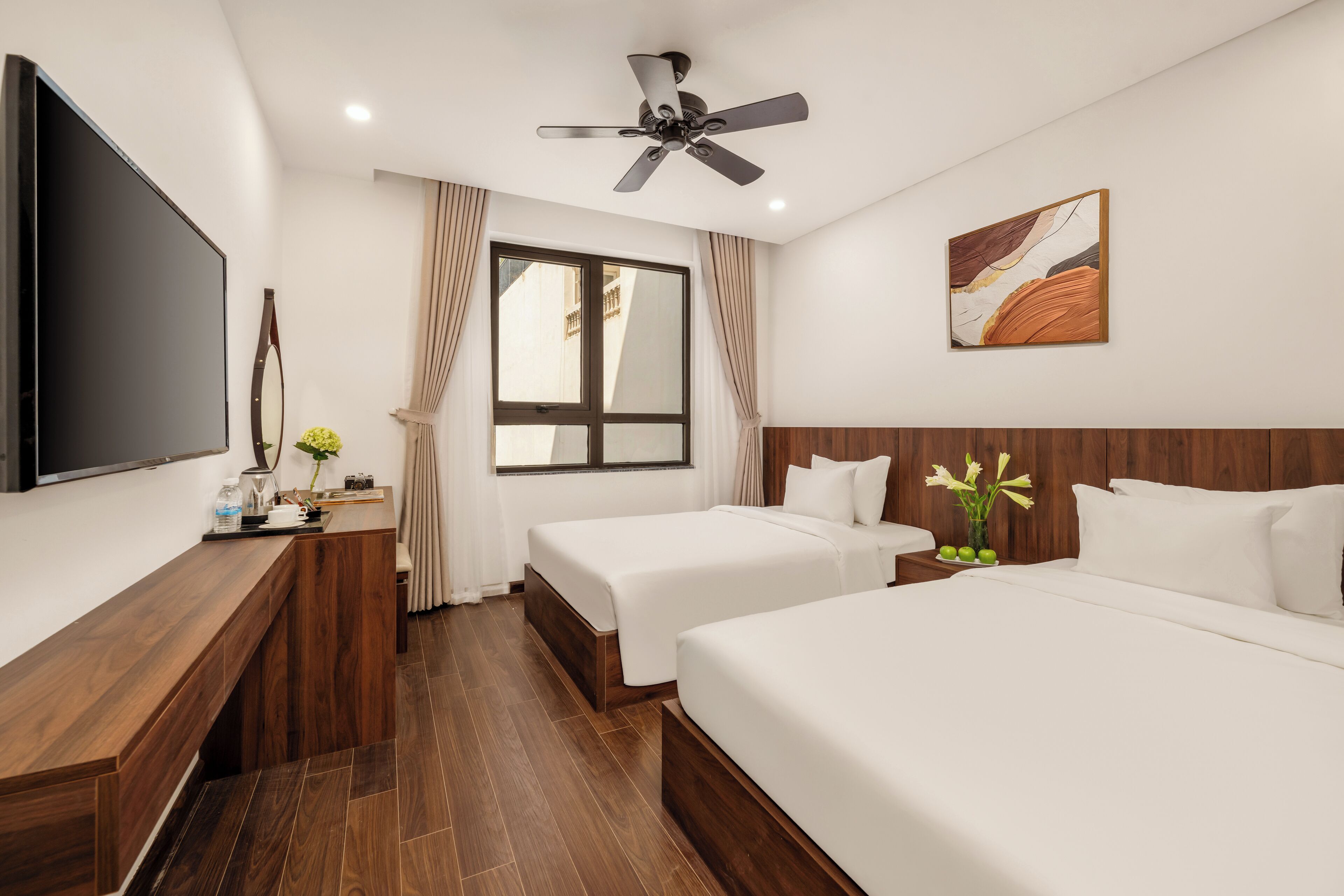 A hotel room with a flat-screen TV, two beds, a desk, a ceiling fan, and a window with curtains.
