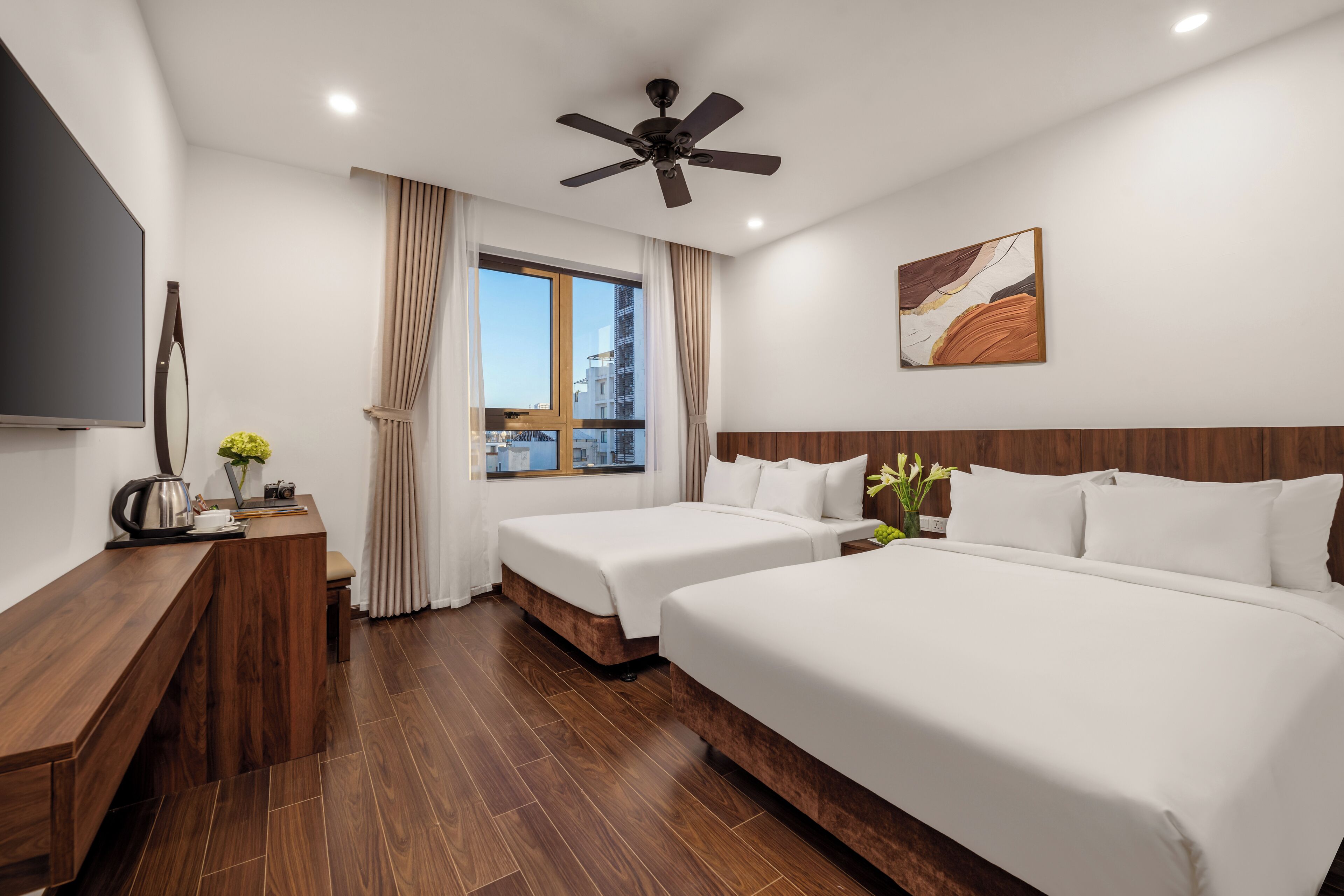A hotel room with two beds, a TV, a wooden desk, a ceiling fan, and a window with curtains.