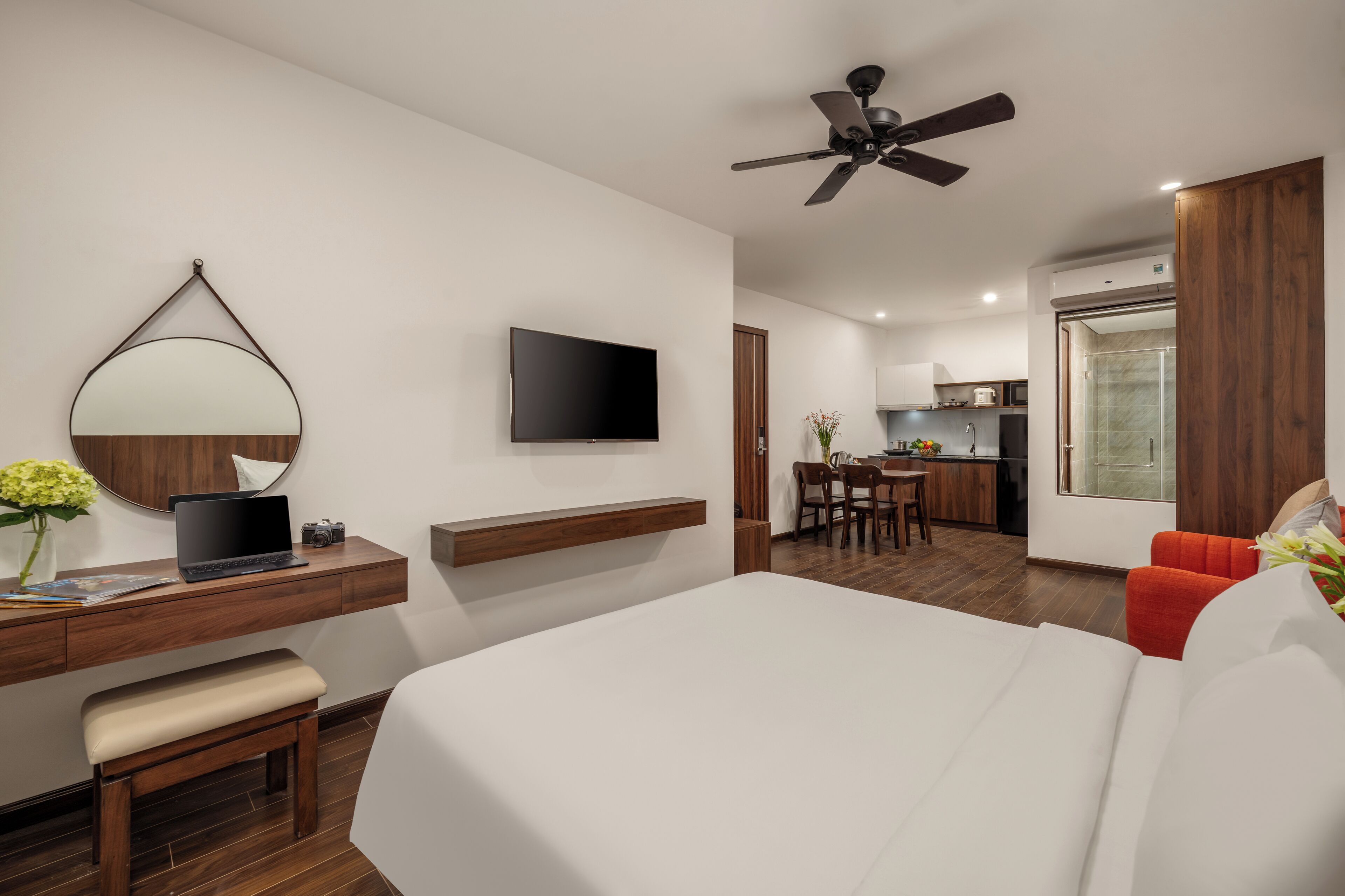Een moderne hotelkamer met een groot bed, een bureau met een laptop, een flatscreen-televisie, een plafondventilator en uitzicht op de keuken en eetruimte.