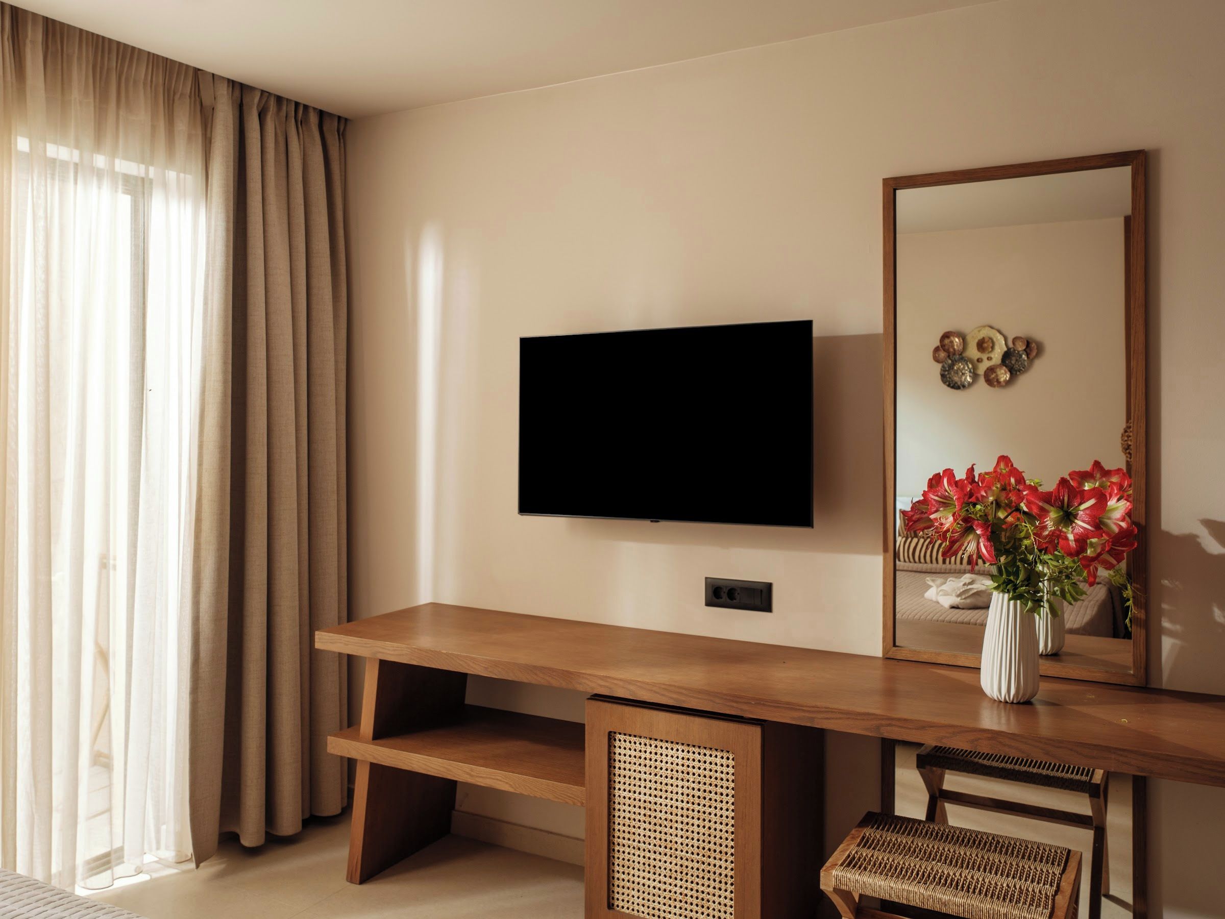 A modern living room with a flat-screen TV mounted on the wall, a wooden coffee table, a wicker chair, and a large mirror reflecting a vase of red flowers.
