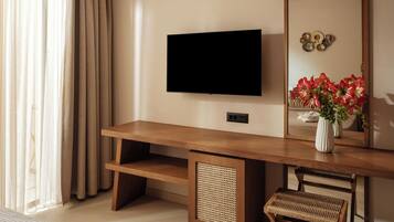 A modern living room with a flat-screen TV mounted on the wall, a wooden coffee table, a wicker chair, and a large mirror reflecting a vase of red flowers.
