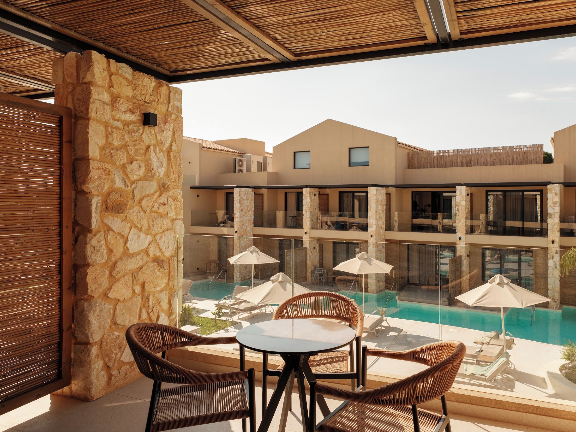 Zona de piscina con mobiliario de mimbre y sombrillas, con vistas a una piscina y un edificio con varios balcones.