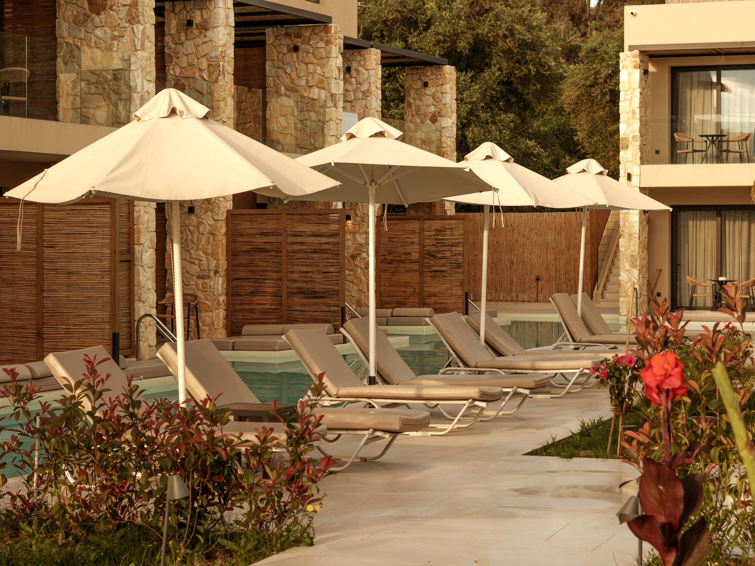 A poolside area with lounge chairs and umbrellas, stone walls, and a wooden fence.