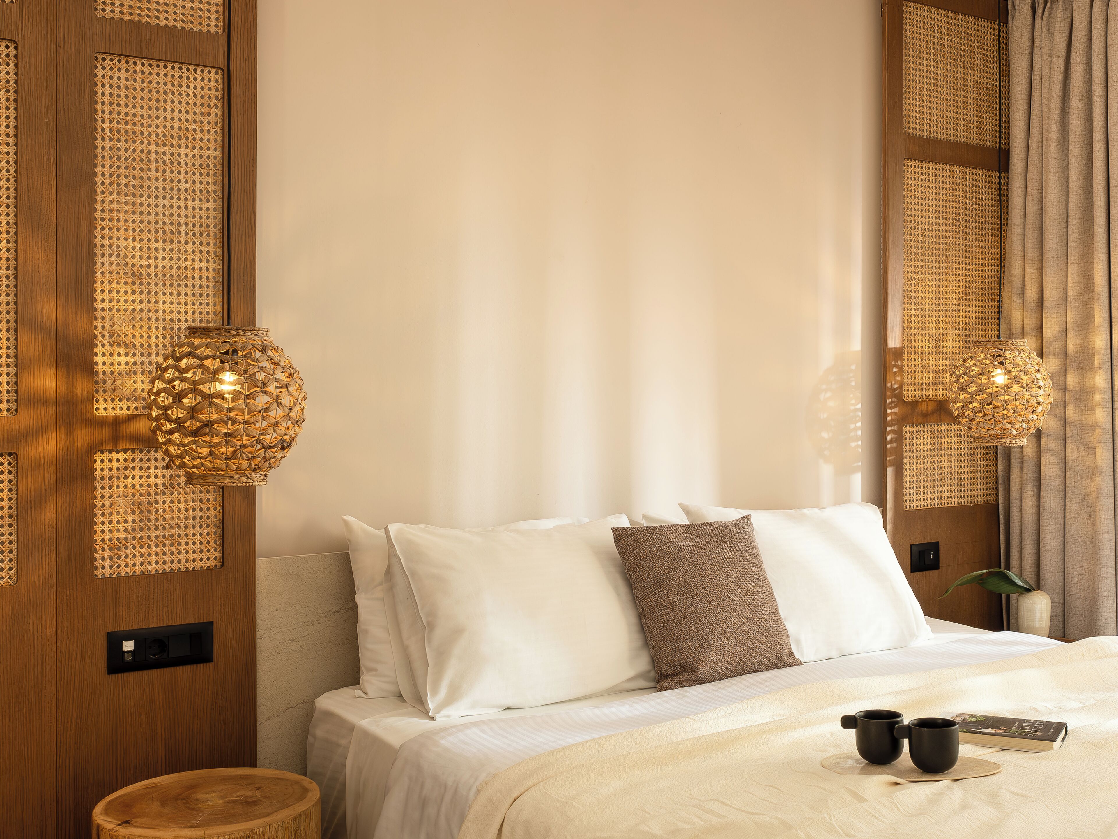 A neatly made bed with white and brown pillows, a bedside table with two black cups and a book, and a woven lamp on the wall.