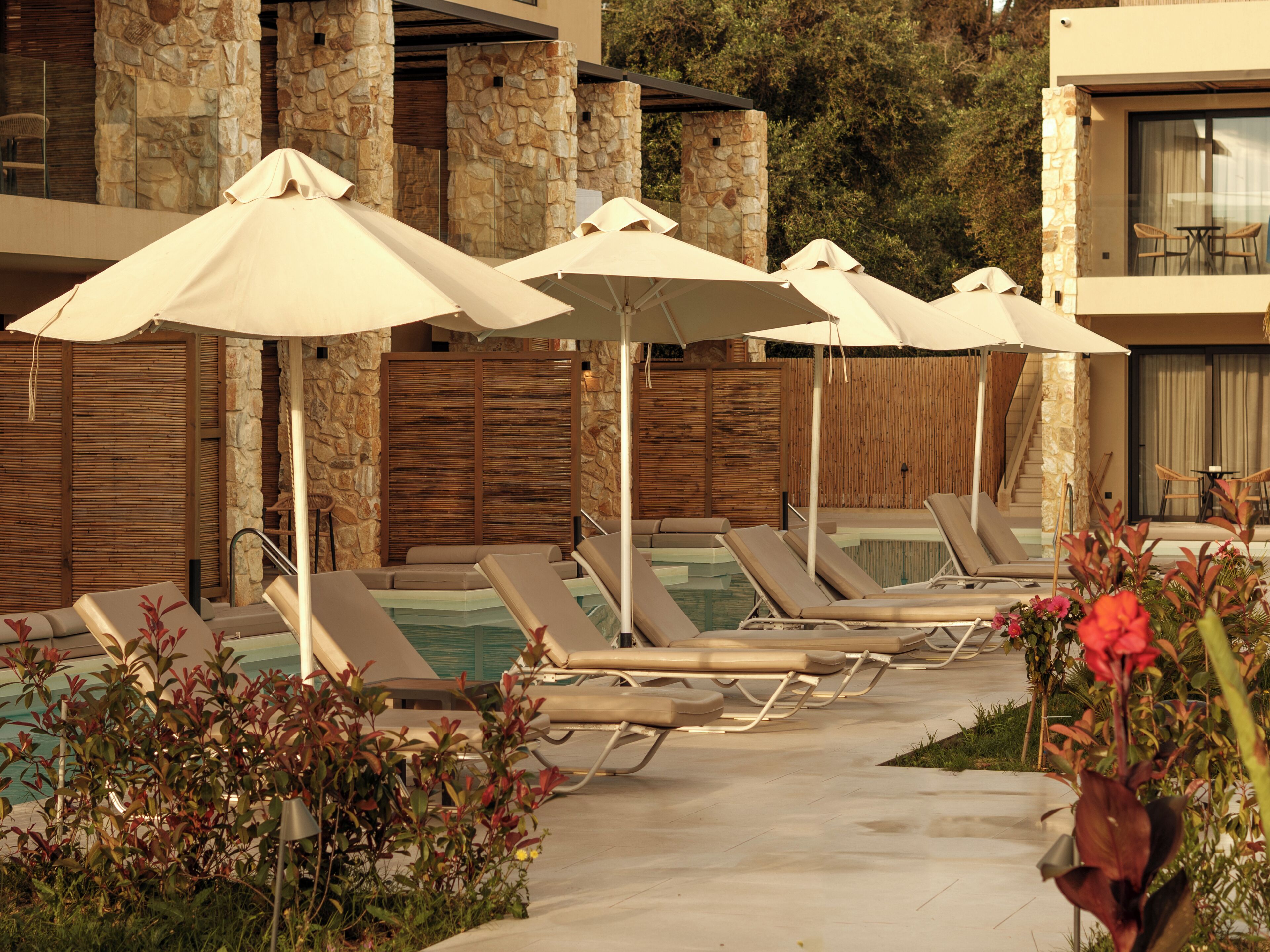 A poolside area with lounge chairs and umbrellas, stone walls, and a wooden fence.