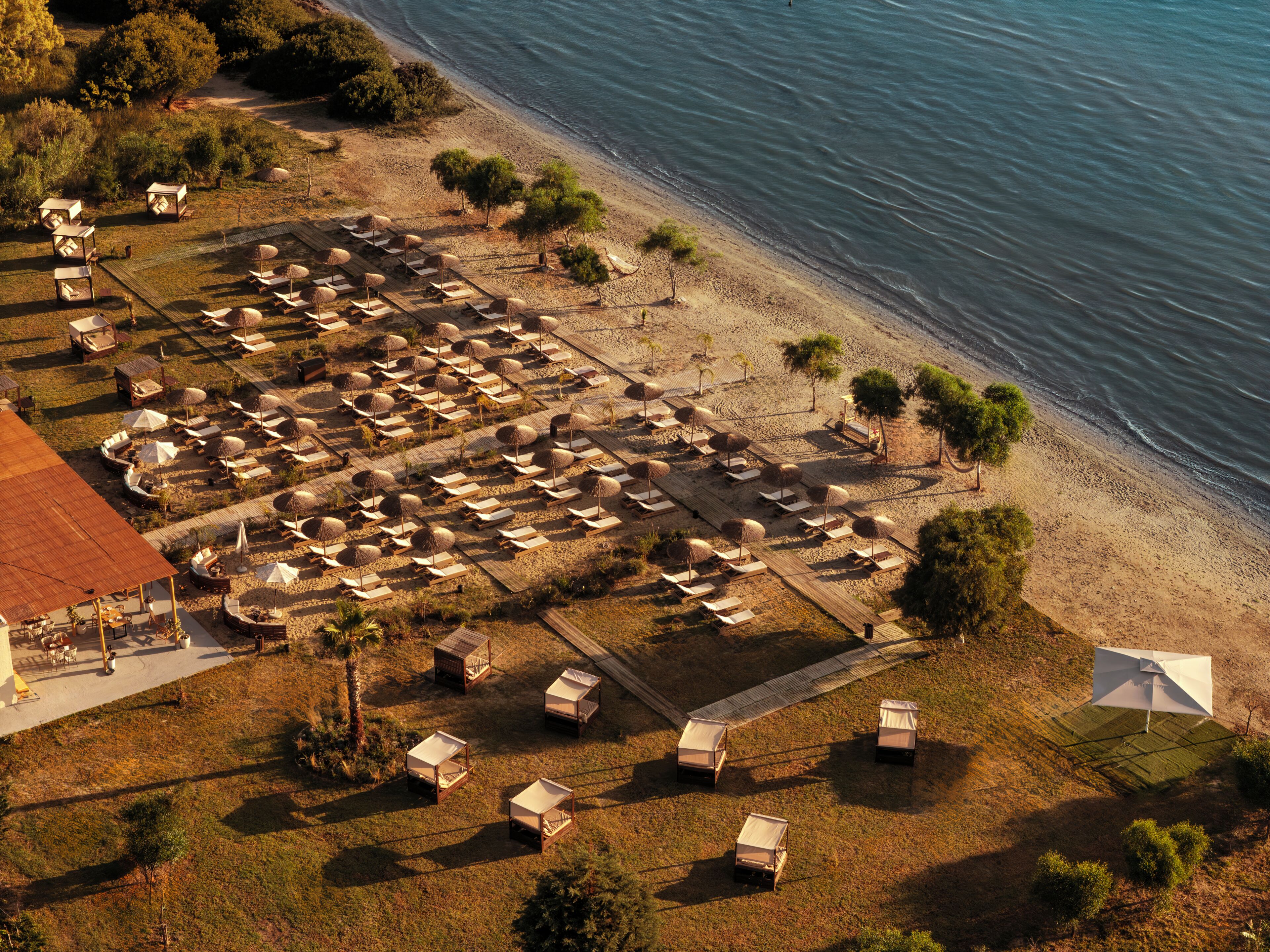 Eine Luftaufnahme eines Strandresorts mit mehreren Bungalows, von denen einige mit Sonnenschirmen versehen sind, auf einer Sandfläche mit einer Holzterrasse.