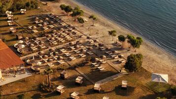 An aerial view of a beach resort with multiple bungalow-style accommodations, some with umbrellas, arranged on a sandy area with a wooden deck.