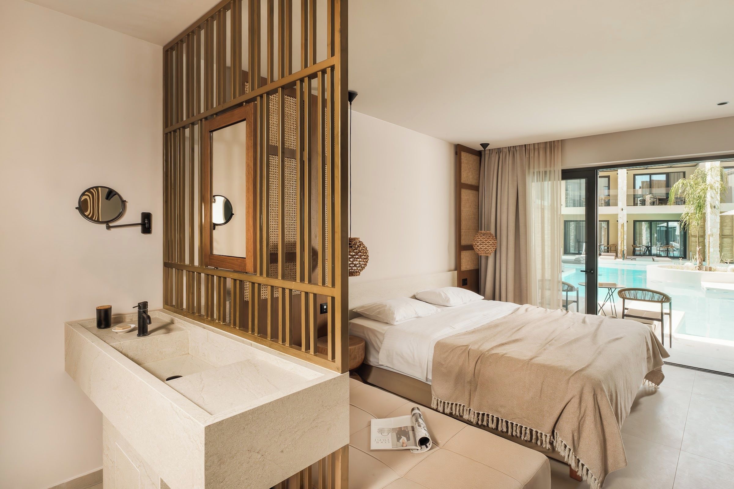 Habitación de hotel con un ventanal grande, una cama con una manta color beige, un espejo redondo y un separador de madera.