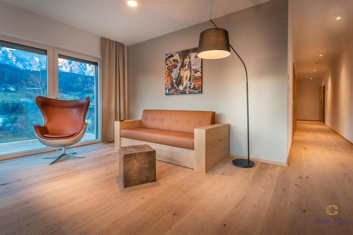 Una sala de estar moderna con un sofá de cuero, una silla original, una lámpara de pie y un amplio ventanal con vistas a la montaña.