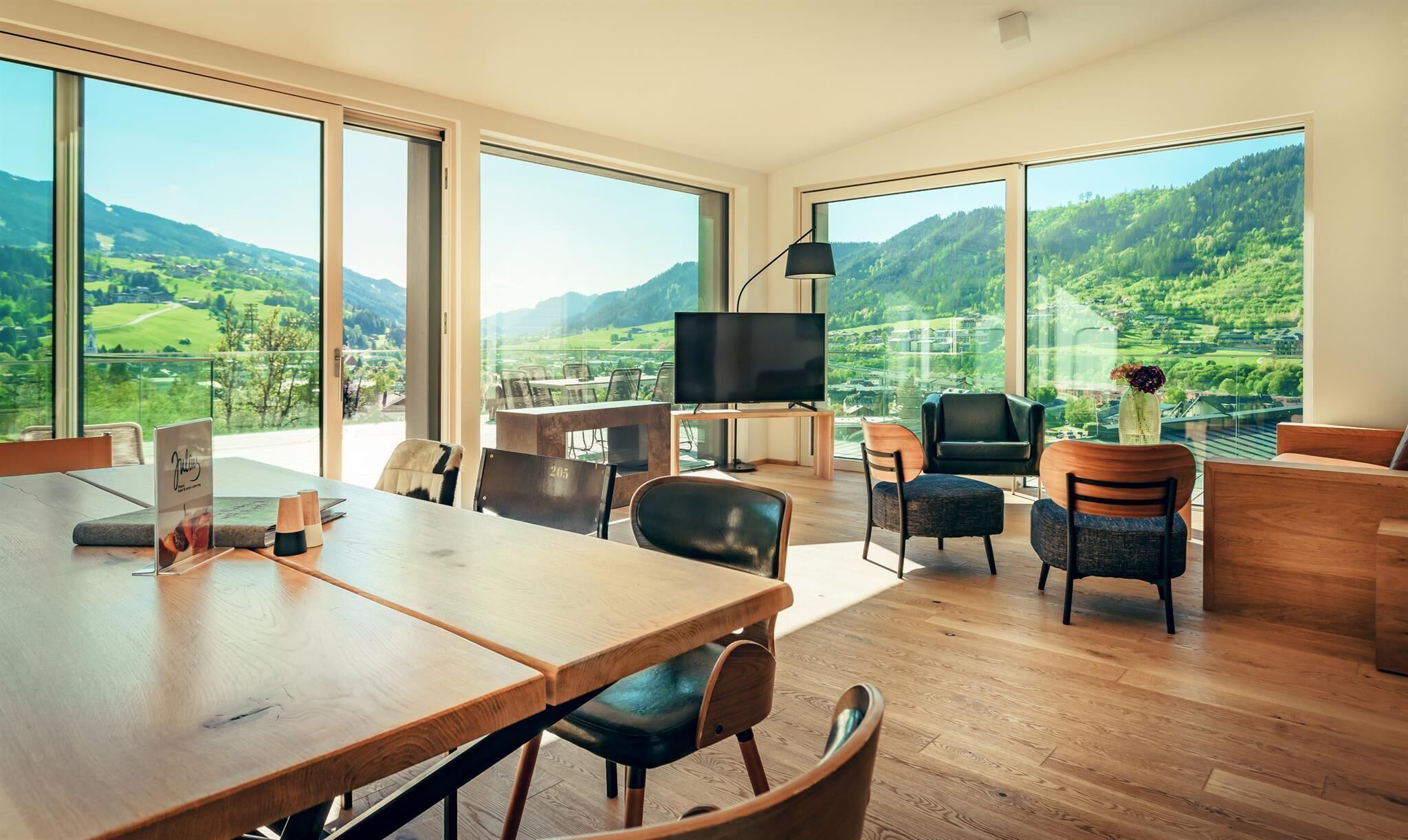 A spacious living area with a large wooden table, comfortable seating, a flat-screen TV, and a scenic mountain view through large windows.