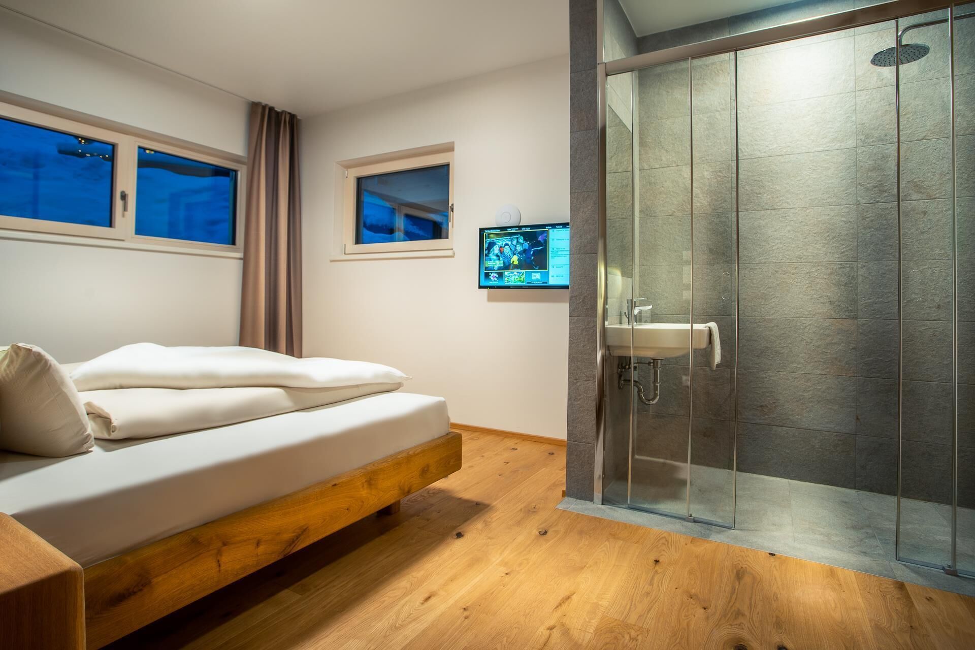 Un dormitorio moderno con una cama de madera, cabina de ducha con mampara de vidrio y un televisor montado en la pared.