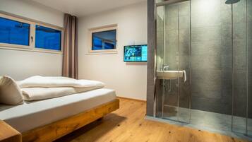 Un dormitorio moderno con una cama de madera, cabina de ducha con mampara de vidrio y un televisor montado en la pared.