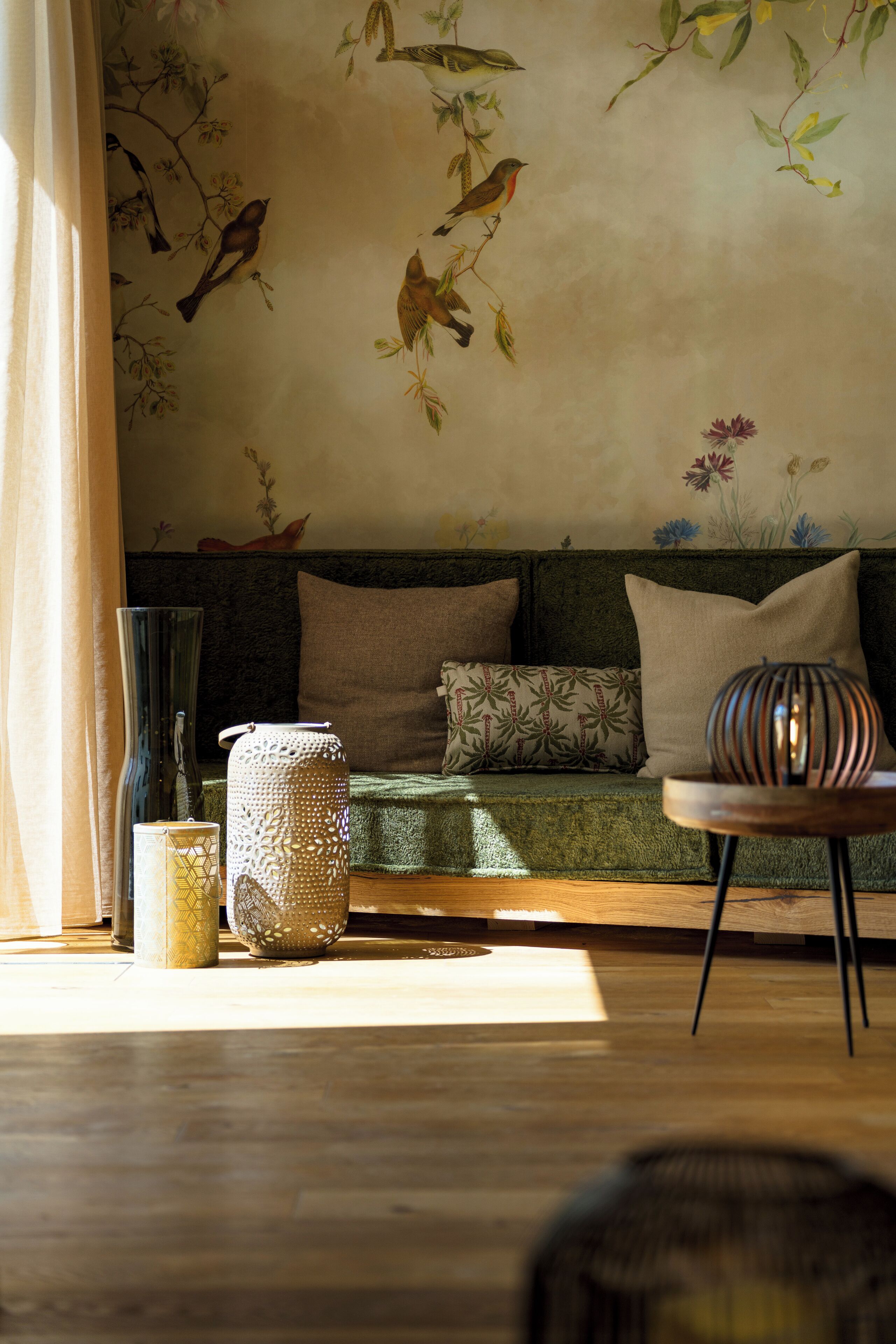 Una sala de estar acogedora con un sofá verde, cojines estampados, un jarrón decorativo y un mural de aves y flores.
