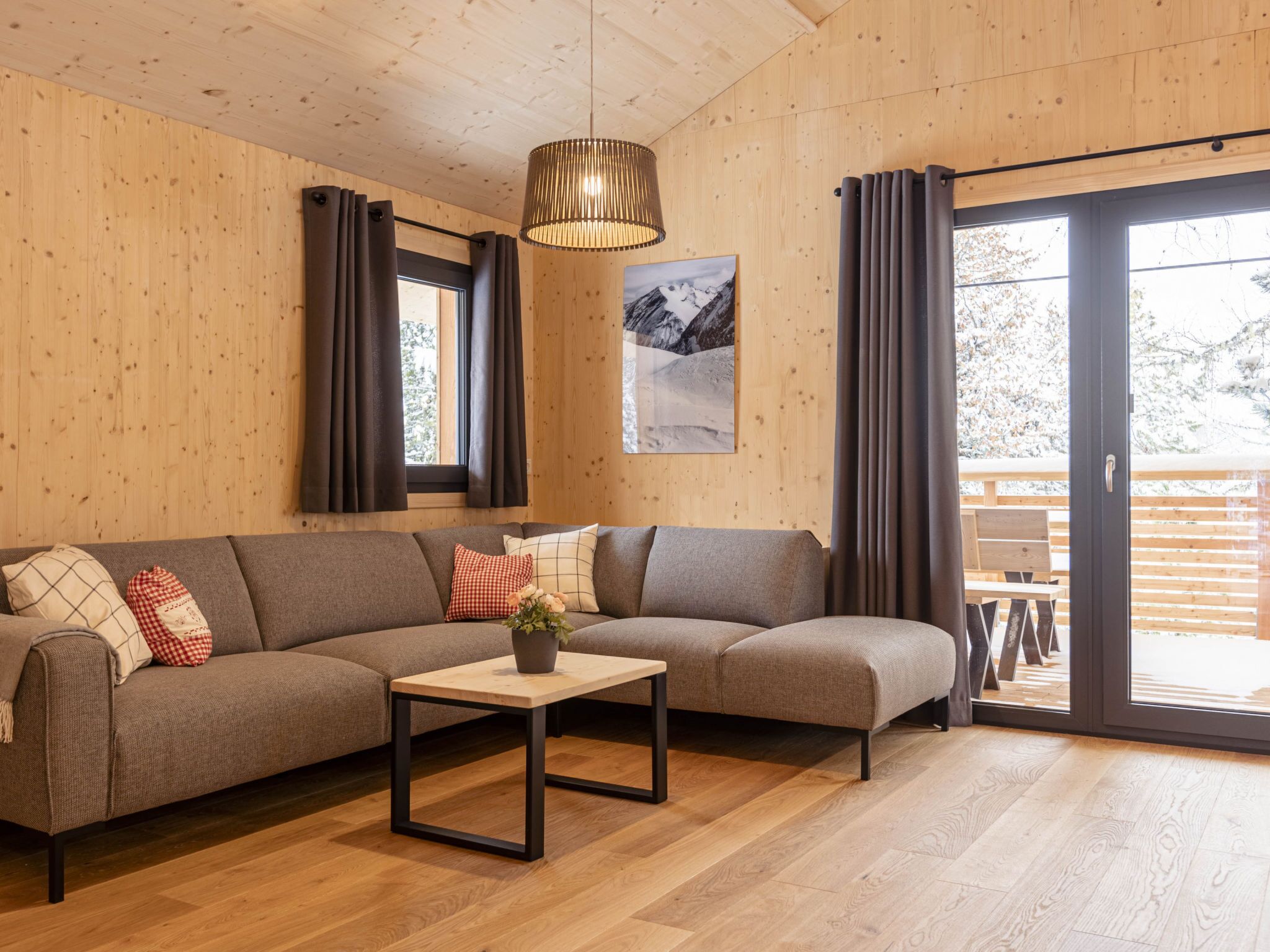 Ein modernes Wohnzimmer mit einer Eckcouch, einem kleinen Tisch mit einer Vase und einem großen Fenster mit Vorhängen.
