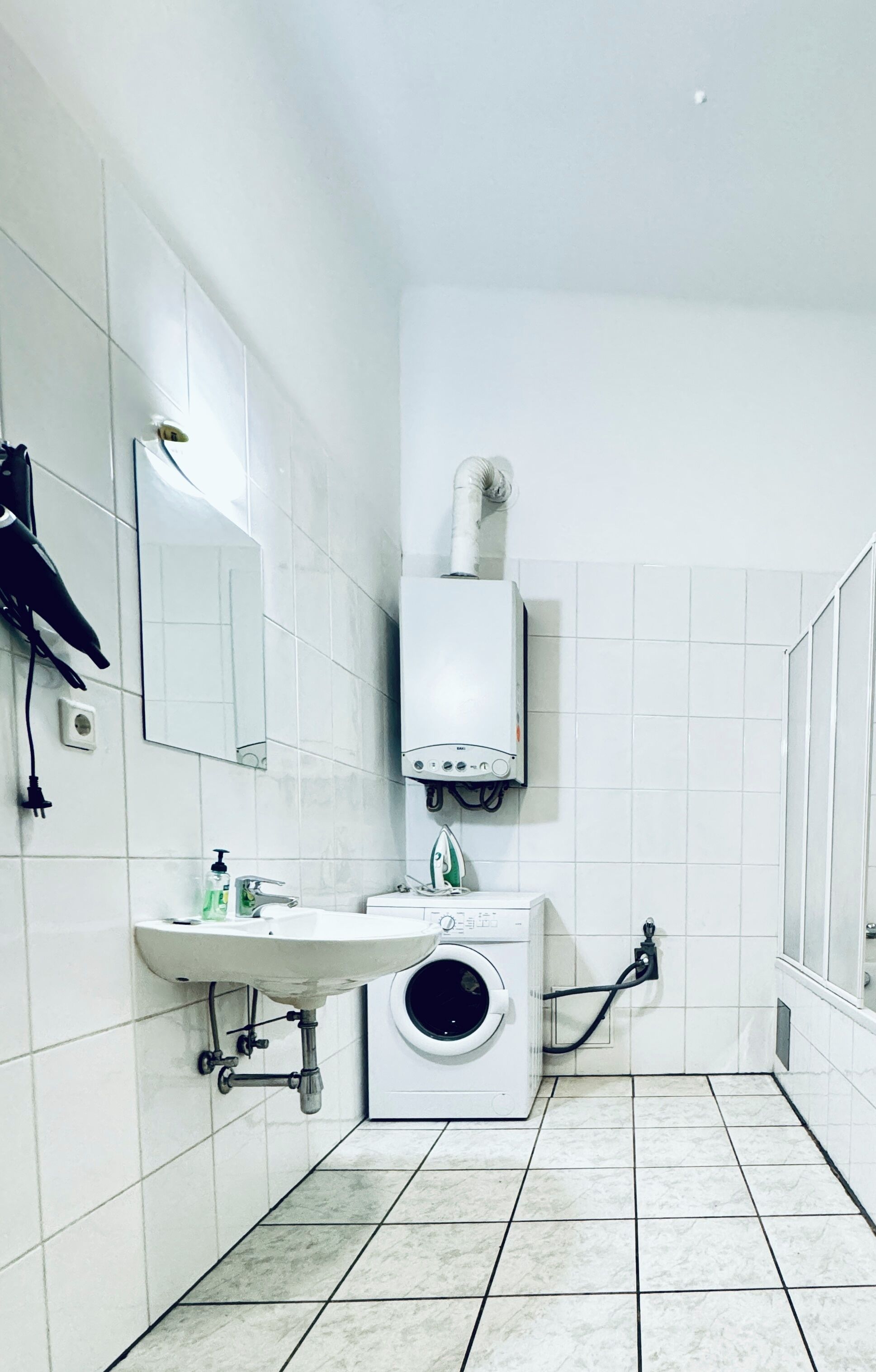 Un baño pequeño y limpio con azulejos blancos, un lavabo, un espejo, un secador de pelo y una lavadora.