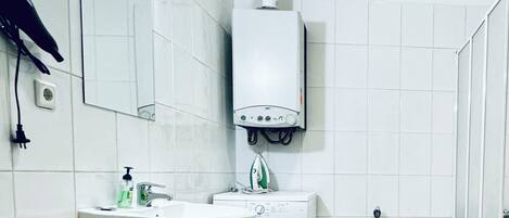 Un baño pequeño y limpio con azulejos blancos, un lavabo, un espejo, un secador de pelo y una lavadora.