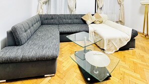 Un salon moderne avec un canapé d’angle gris, une table basse en verre et un lit recouvert d’une couette blanche.