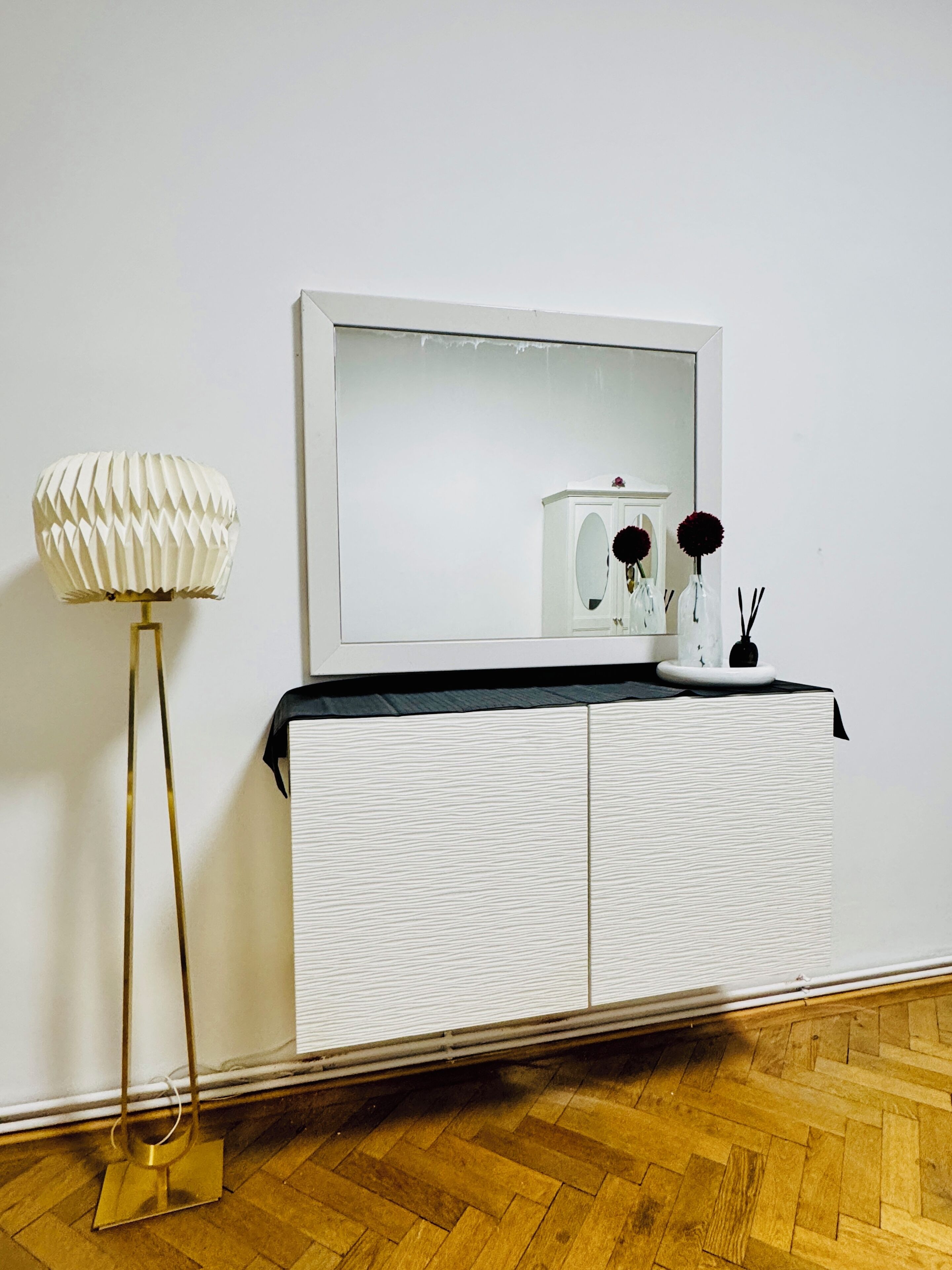 Un meuble blanc avec un miroir au-dessus, un lampadaire doré et une table noire agrémentée d’objets décoratifs.