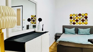 Une chambre moderne avec un grand miroir, une table de chevet avec une lampe, un lit avec une literie bleue et une œuvre d’art murale.