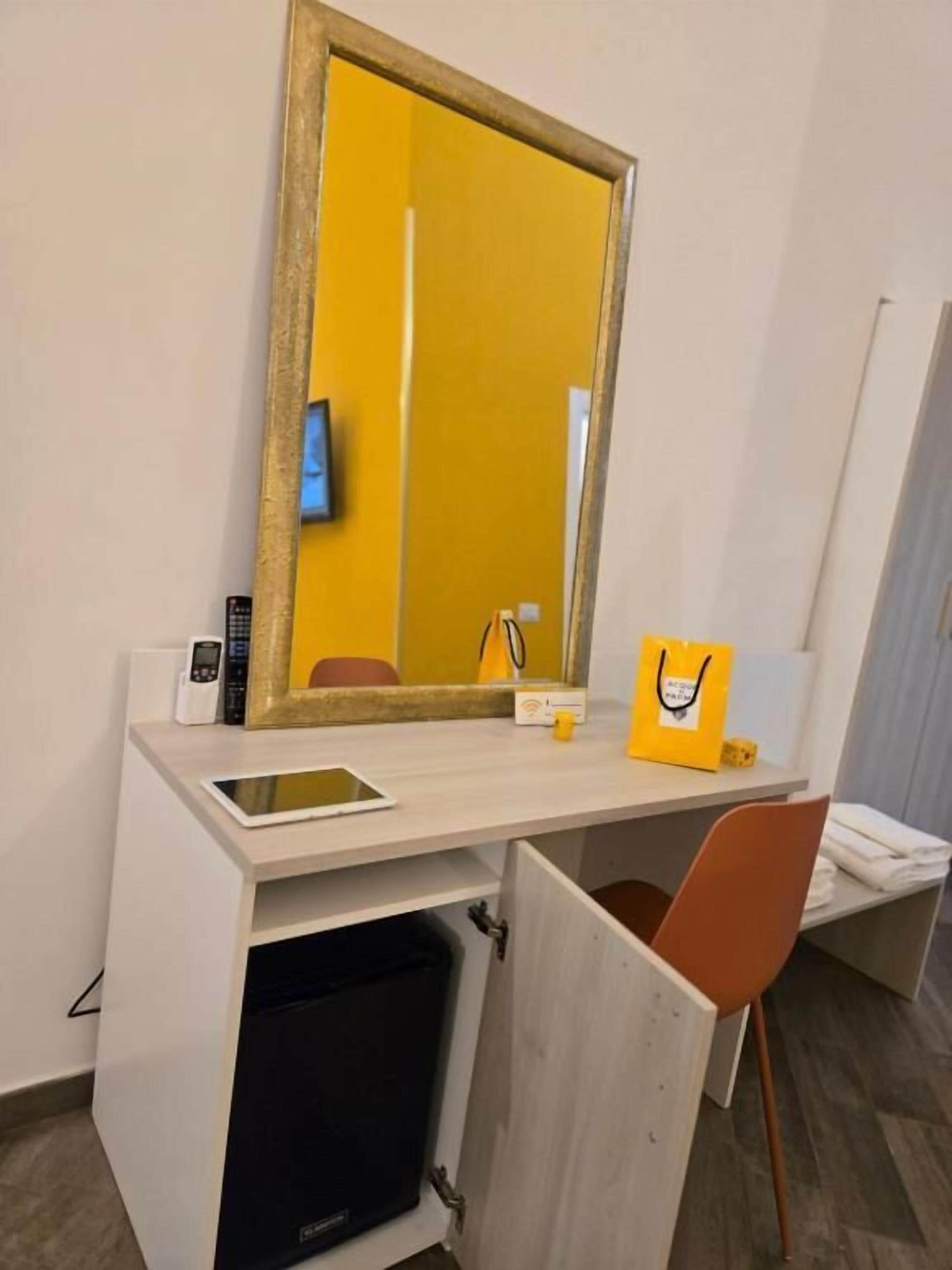 Een hotelkamer met een bureau, een spiegel, een stoel, een klein zwart apparaat en een gele tas op het bureau.