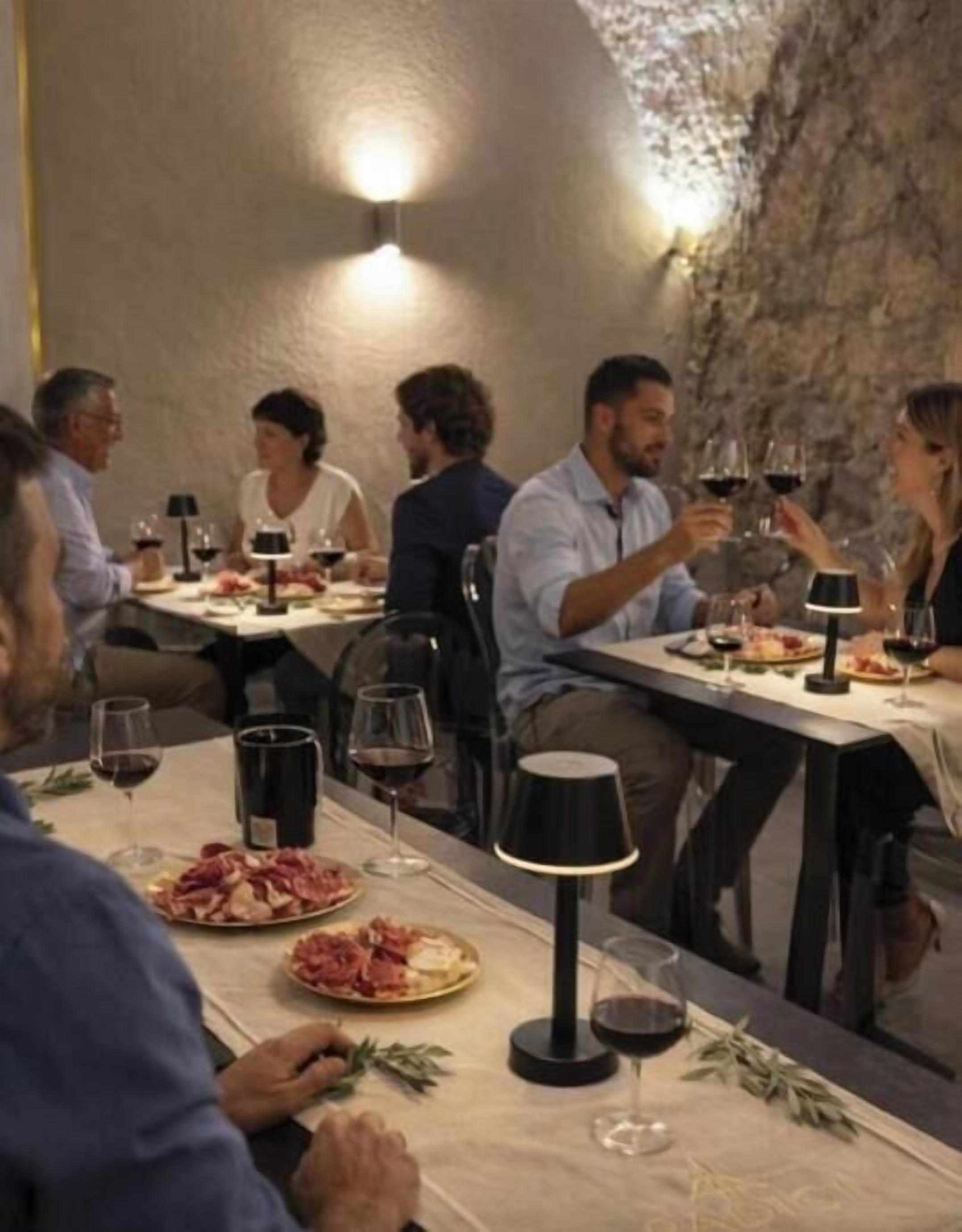 Un grupo de personas cenando en una mesa, con copas de vino, platos de comida y una lámpara.