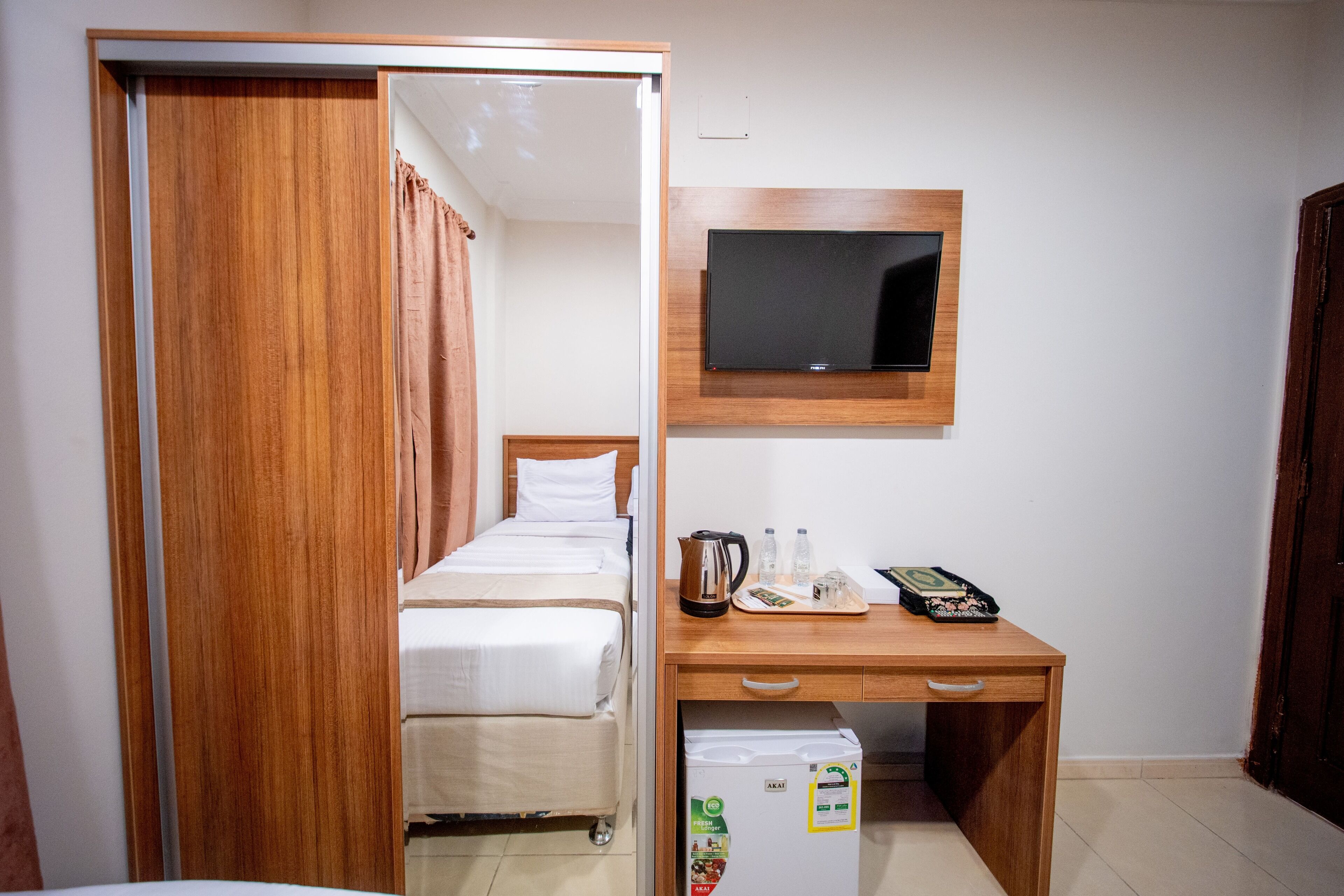 A hotel room with a wooden wardrobe, a bed with white bedding, a desk with a television, and a mini-fridge.