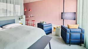 Une chambre d’hôtel moderne, équipée d’un lit, d’un bureau, de deux canapés bleus, d’une télévision et d’une grande fenêtre avec des stores.