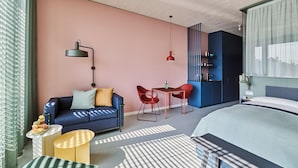 Une chambre d’hôtel moderne avec un canapé bleu, une petite table à manger, des chaises rouges, un lit et un coin cuisine.