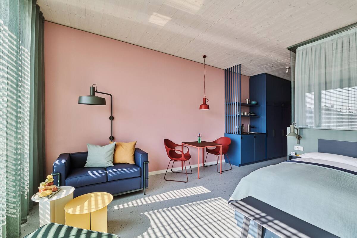 A modern hotel room with a blue sofa, a small dining table, red chairs, a bed, and a kitchenette area.