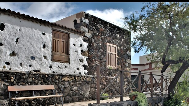 A stone building with a white exterior and wooden shutters, a bench, and a stone wall with greenery. - Vacation Studio 'Tagoro' with Wi-Fi (Guía de Isora)