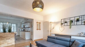 Een moderne woonkamer met een grijze modulaire bank, een houten salontafel met een rode vaas en fles, en een salontafel met een glazen blad. In de achtergrond is een keuken met witte kasten en een hanglamp te zien.