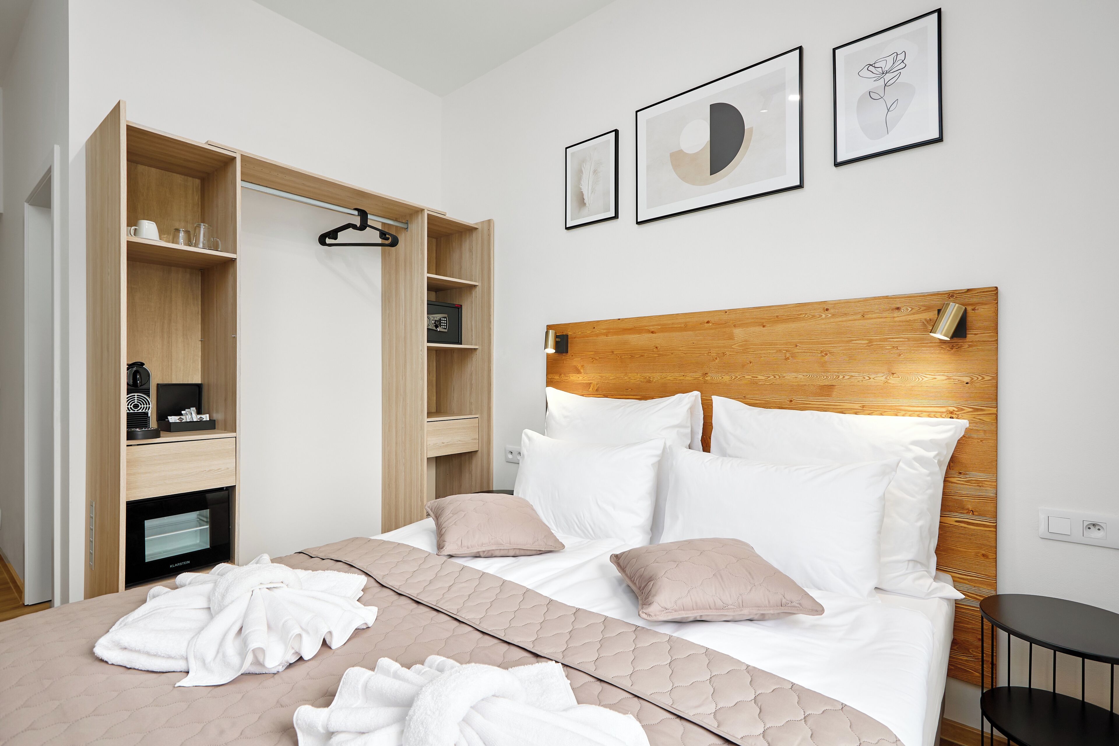 Ein Hotelzimmer mit einem hölzernen Kopfende, einem Bett mit weißer und beiger Bettwäsche, einem Holzschrank mit Regalen und einem kleinen schwarzen Tisch.