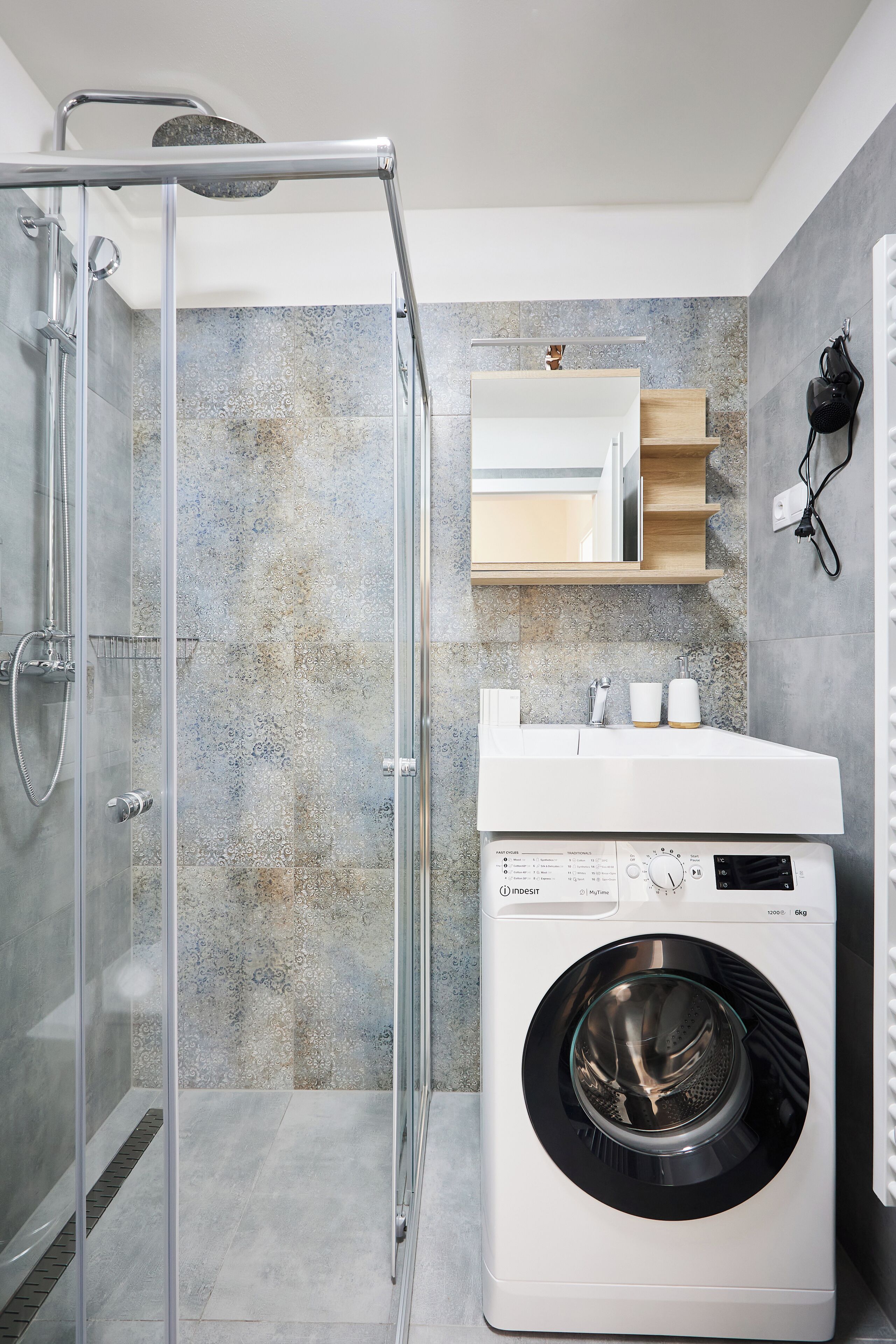 A modern bathroom with a glass shower enclosure, a built-in washing machine, and a mirror above the sink.