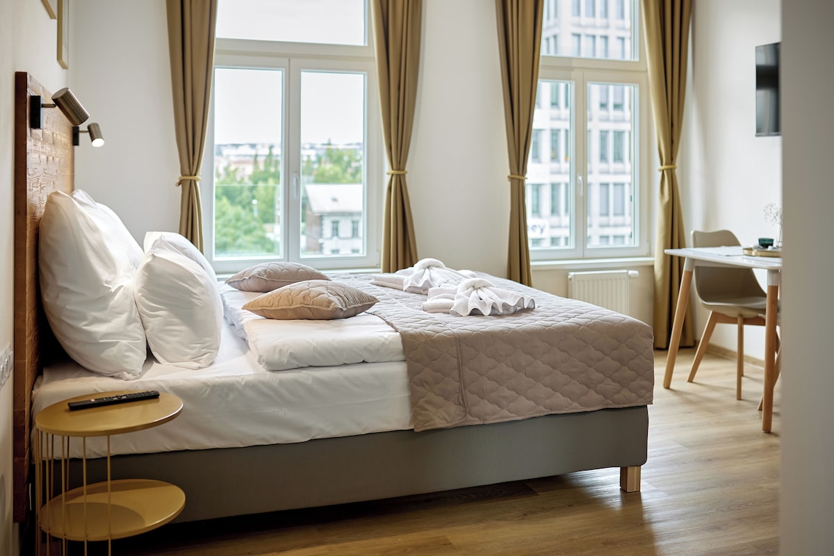 A neatly made bed with pillows, a bedside table with a remote control, and a view of the city through large windows with curtains.