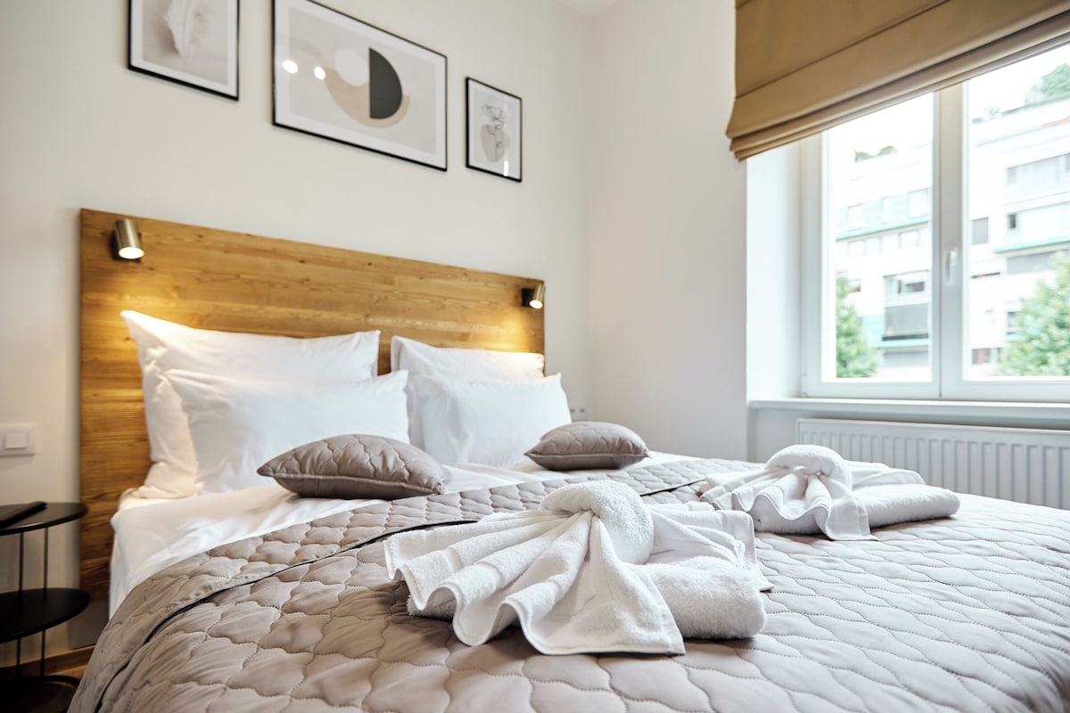 A neatly made bed with a quilted cover, pillows, and folded towels, a wooden headboard, and framed artwork on the wall.