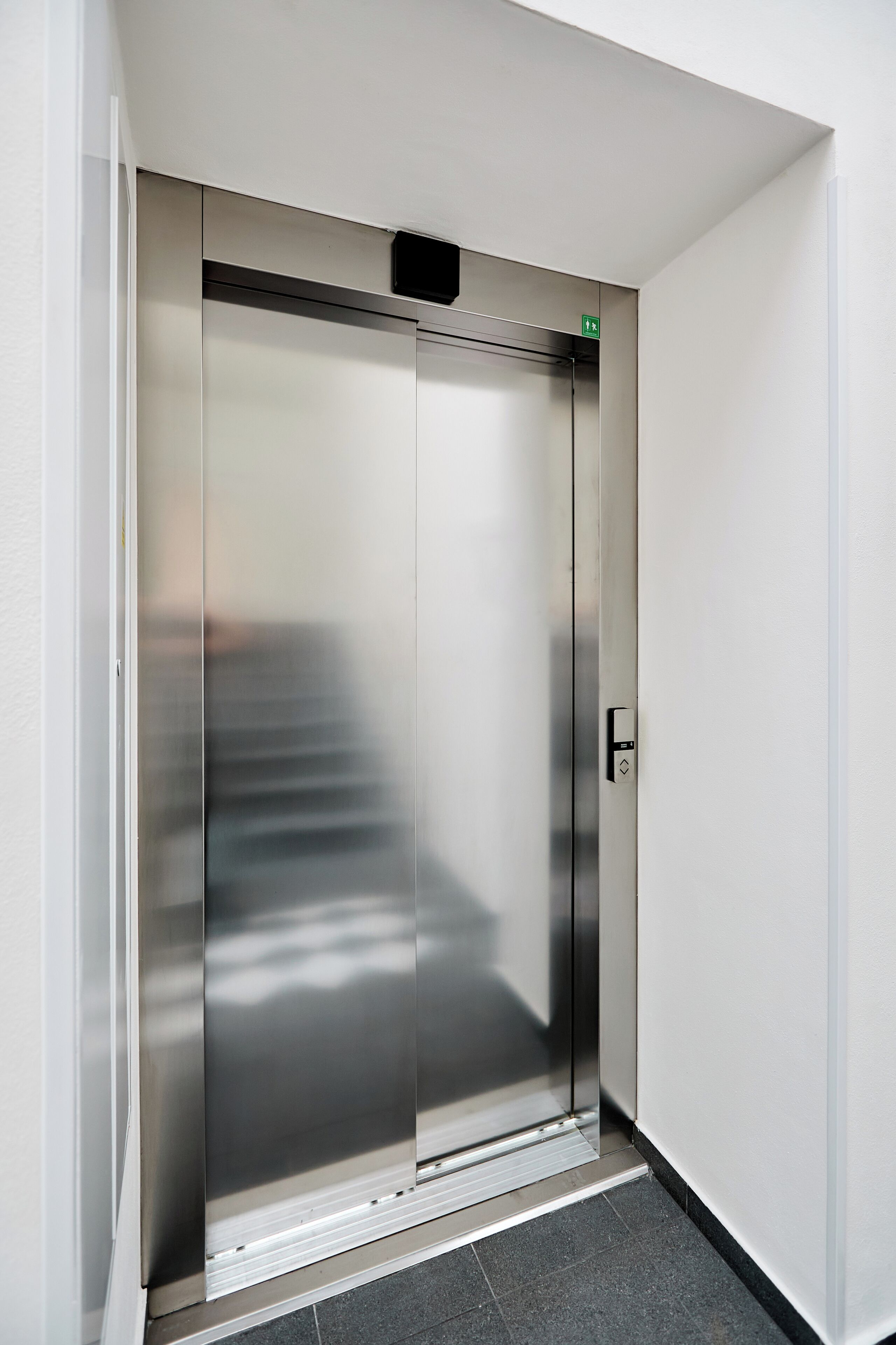 A modern elevator with a metallic finish, a green emergency exit sign, and a control panel on the right side.