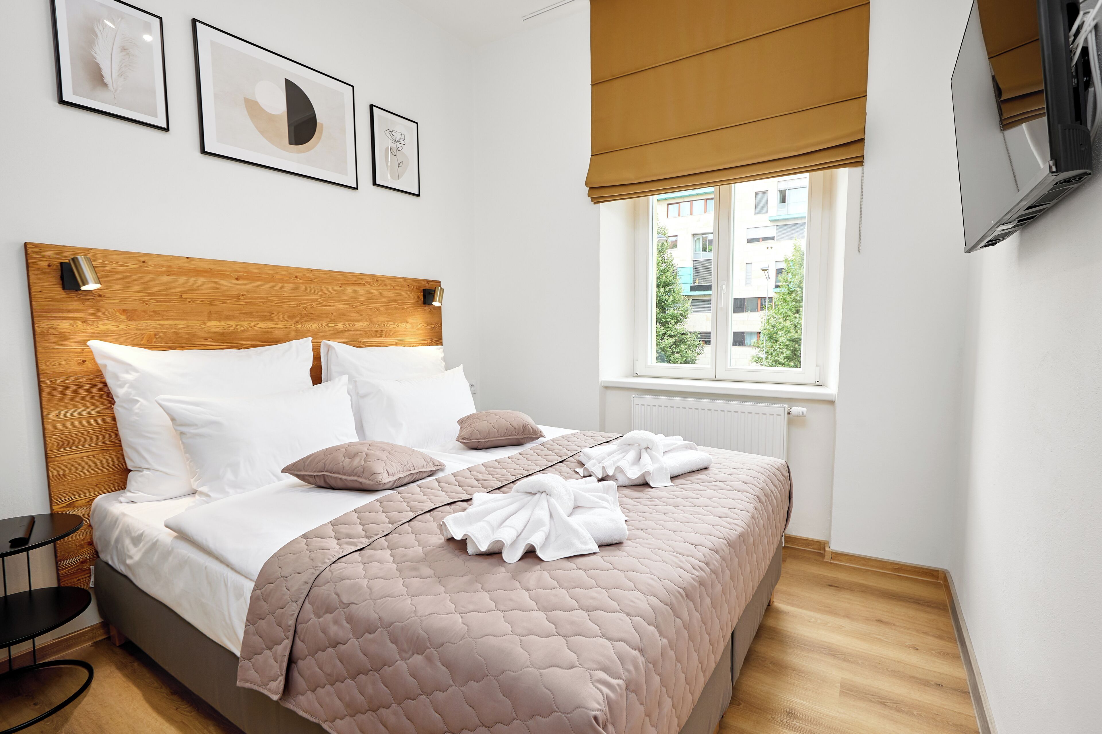 A neatly made bed with a wooden headboard, white and beige pillows, and a pink quilted bedspread. There are two framed artworks on the wall above the bed, and a window with a view of buildings and trees.