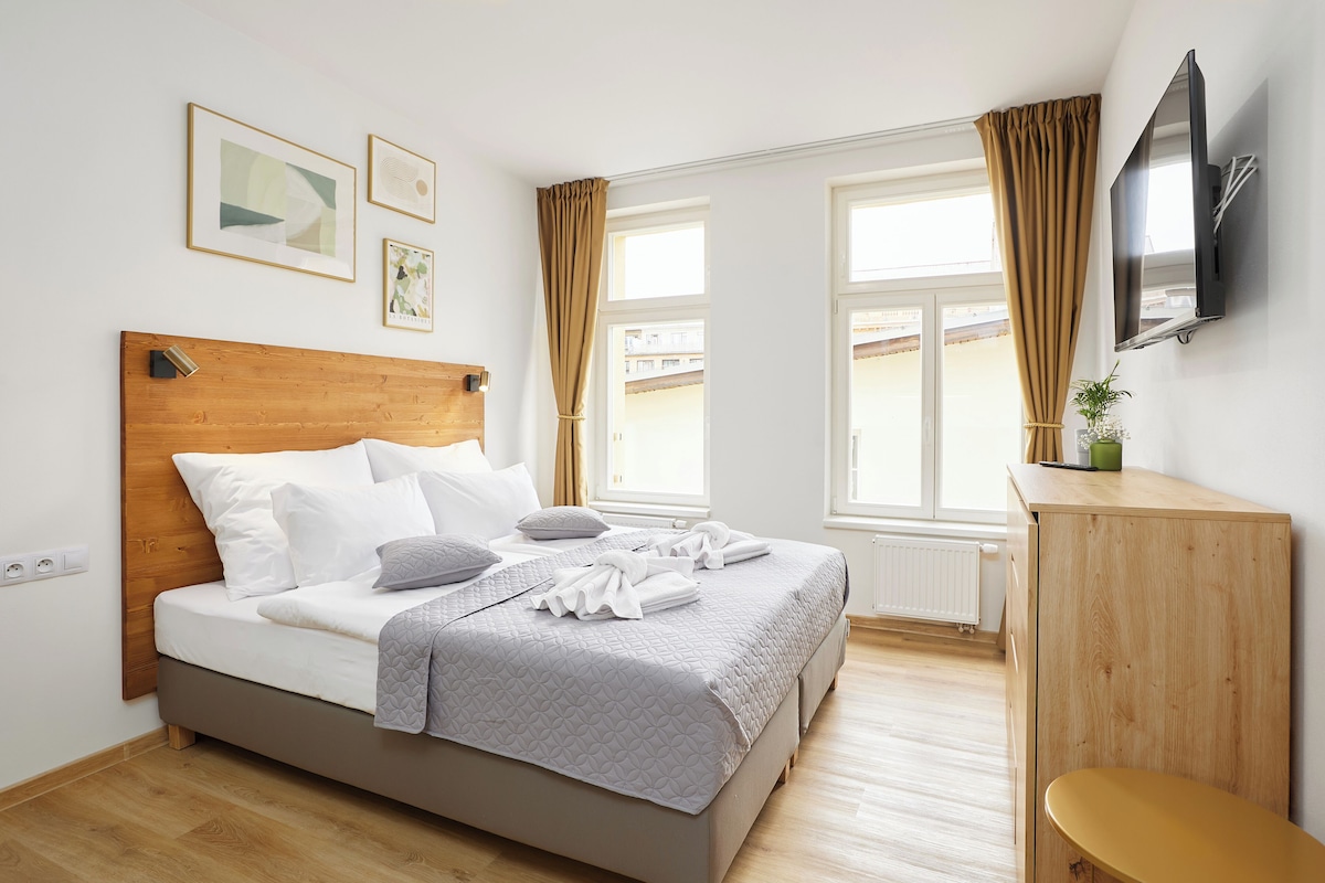 A neatly arranged bedroom with a bed, a wooden headboard, a bedside table, a television, and a window with curtains.