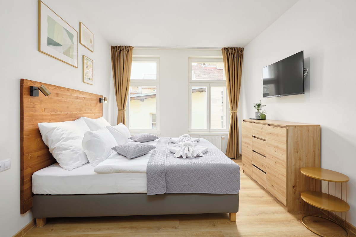 A modern bedroom with a wooden headboard, a bed with white bedding, a grey quilted blanket, a wooden dresser, a flat-screen TV, and a window with curtains.