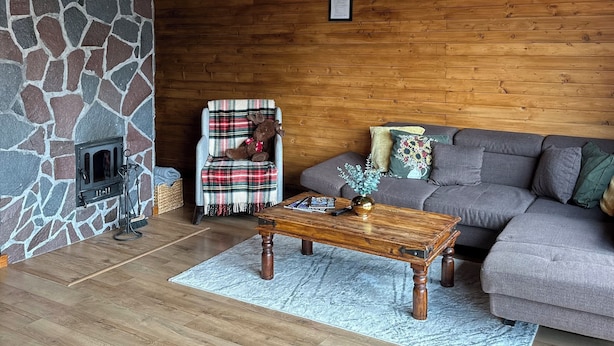 Un salon chaleureux avec une cheminée en pierre, des poutres apparentes en bois au plafond, un fauteuil à carreaux avec un ours en peluche, un canapé gris et une table basse en bois.