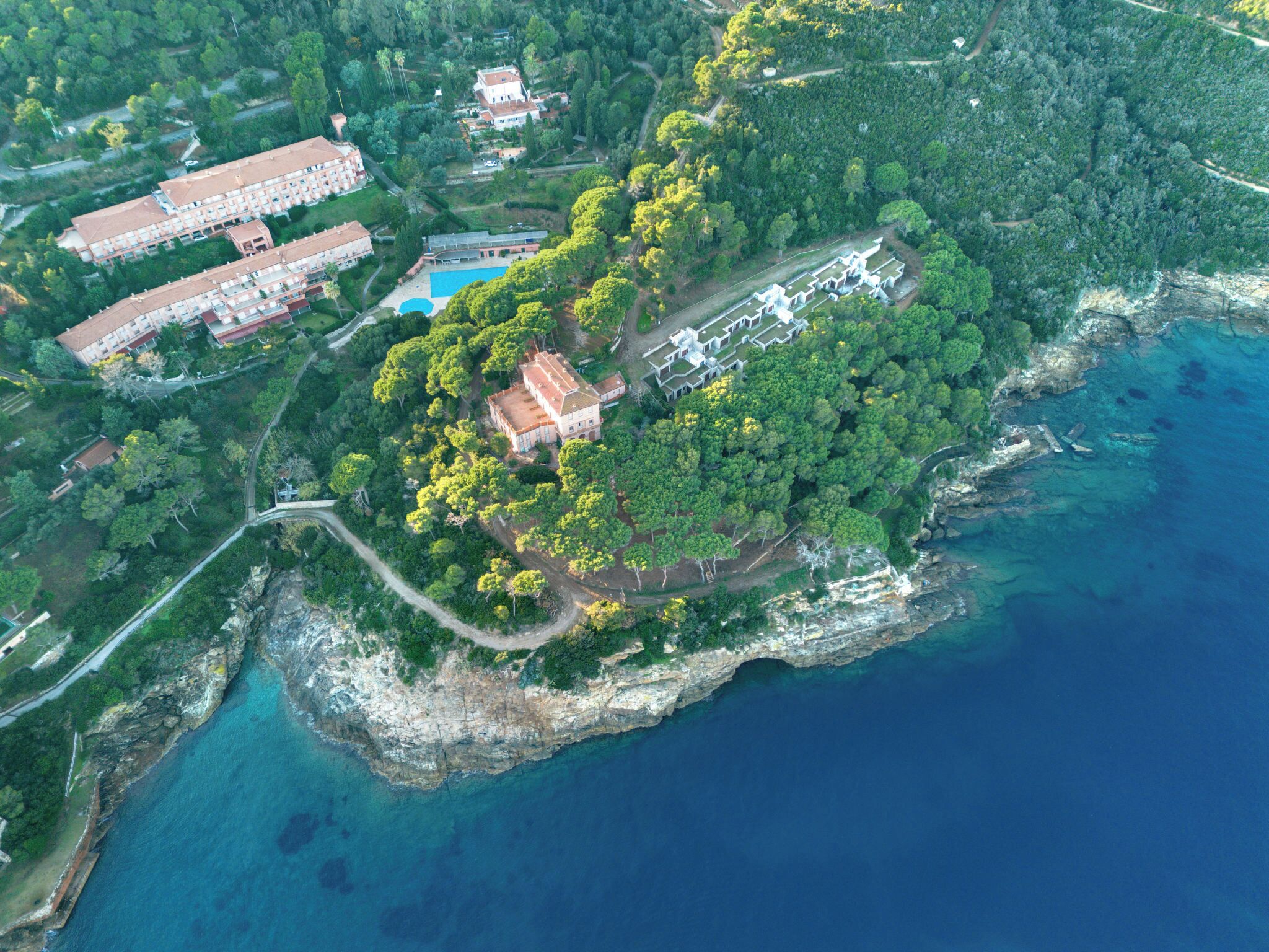 An aerial view of a coastal property with a large building, a swimming pool, and dense greenery.