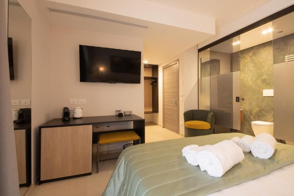 A modern hotel room with a bed, a desk, a chair, a television, and a bathroom with a glass shower enclosure.