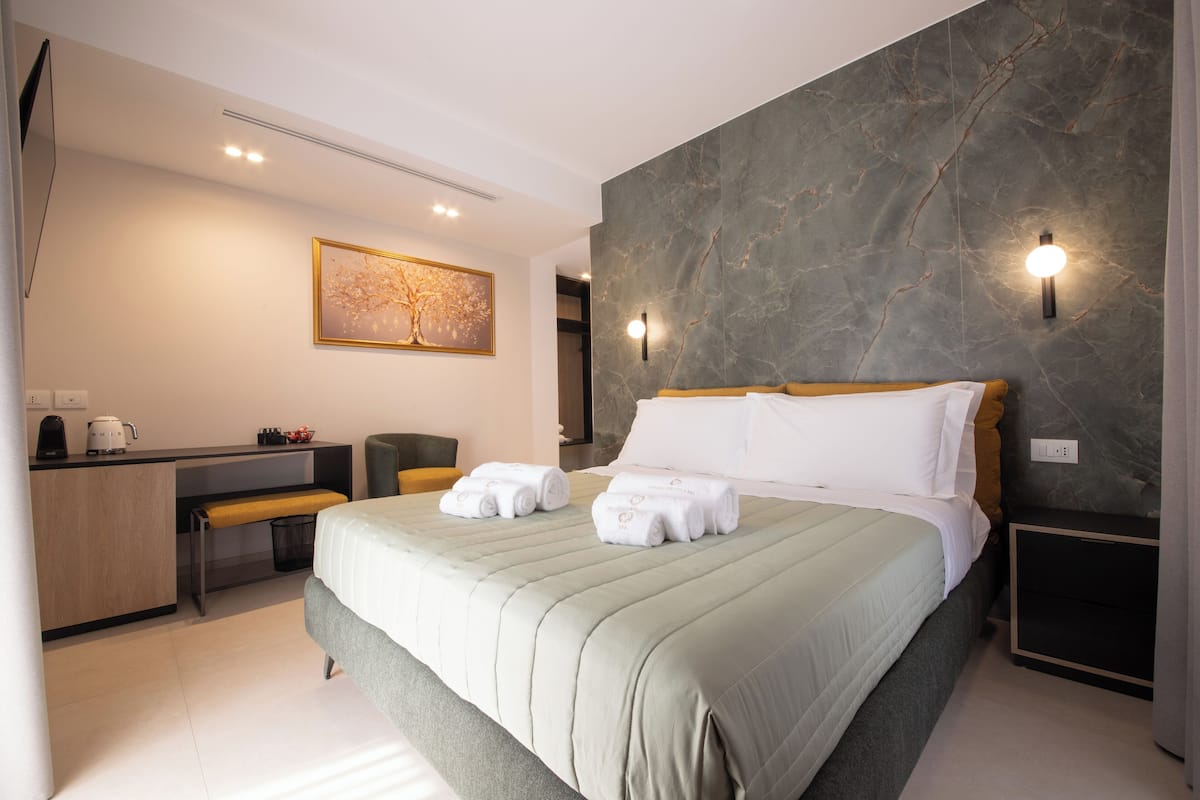 A modern hotel room with a large bed, a desk with a chair, a bench, and a wall-mounted artwork.