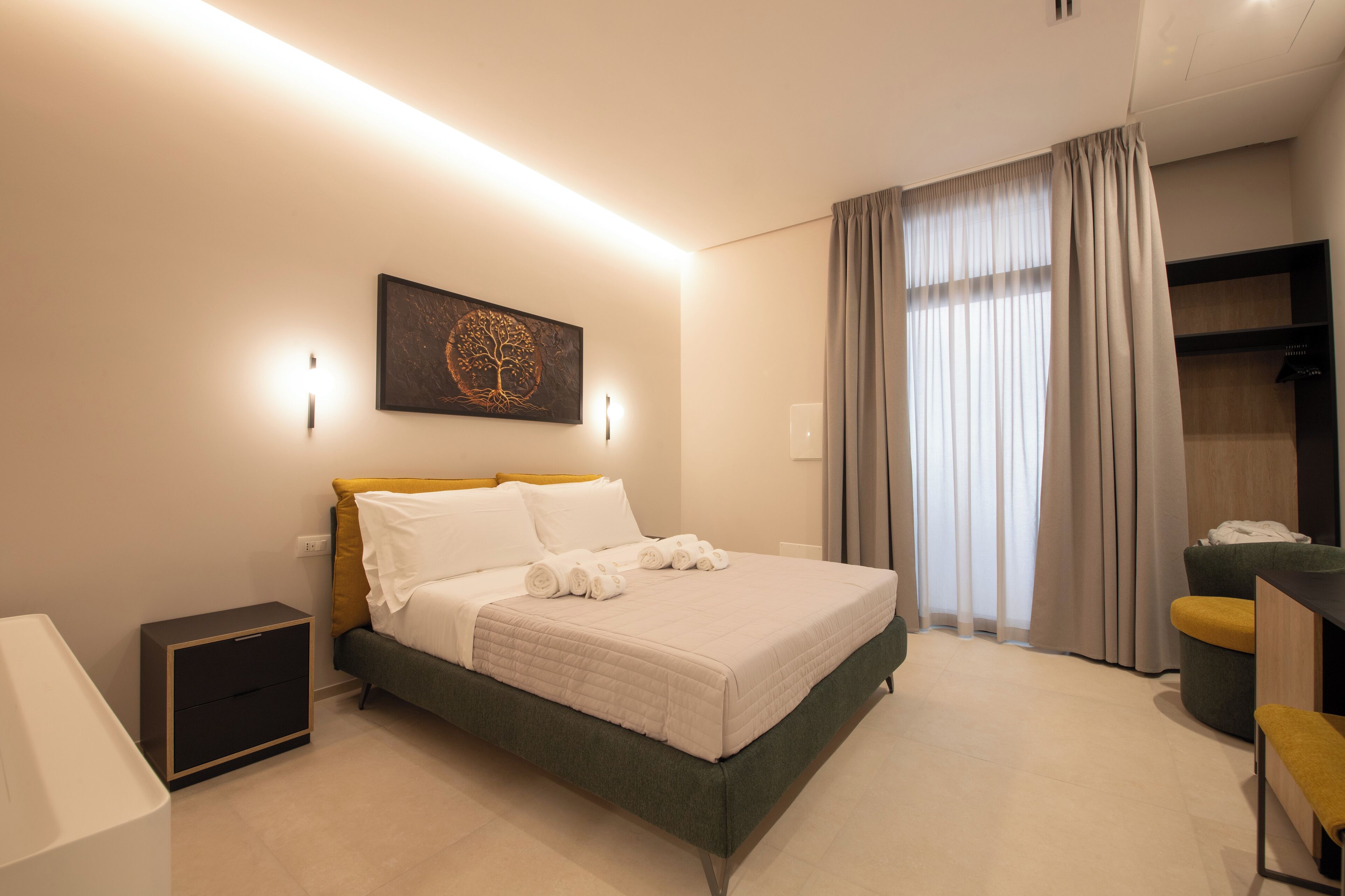 A modern hotel room with a neatly made bed, a bedside table, a desk with a chair, and a large window with curtains.