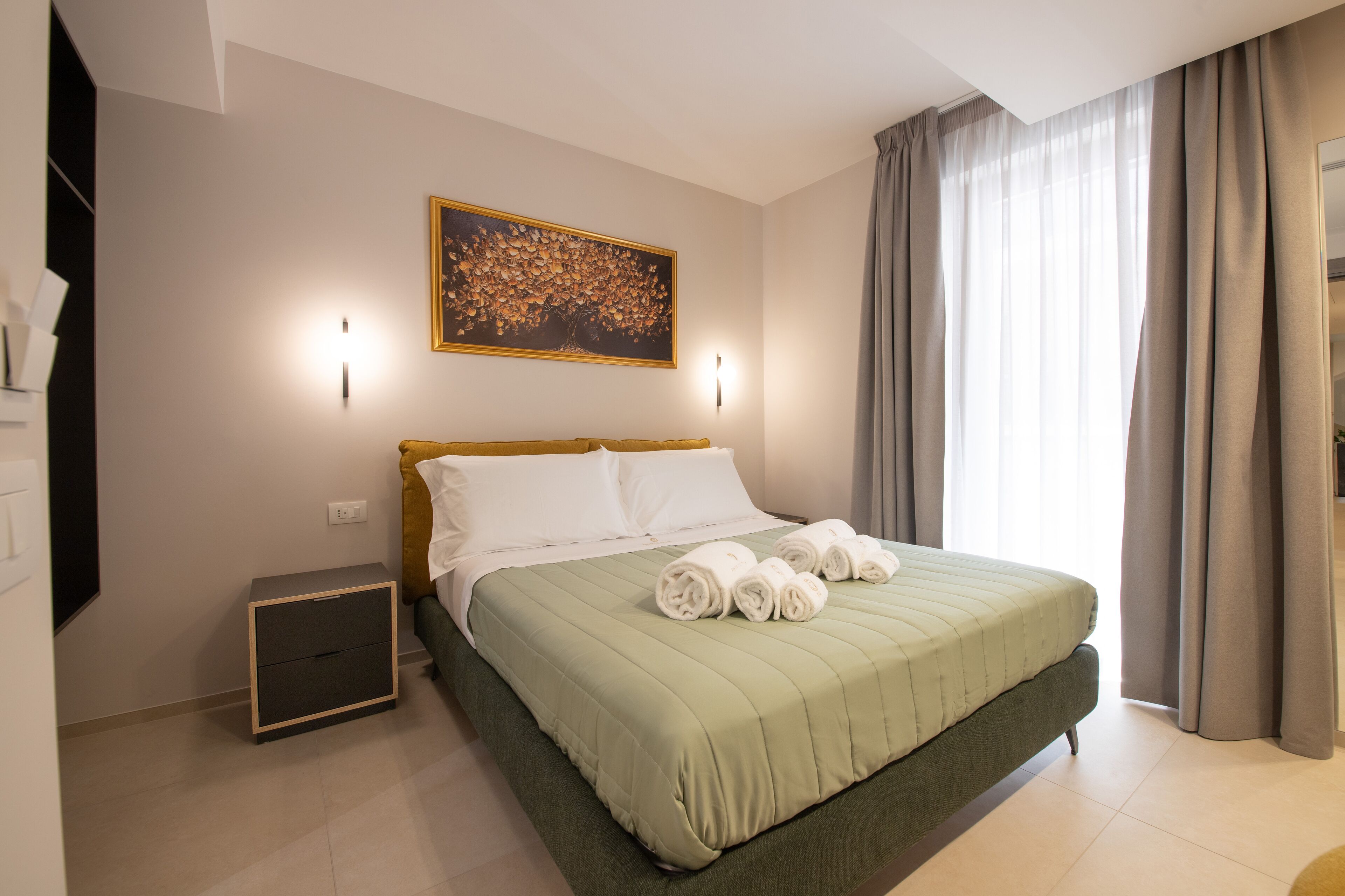 A neatly made bed with a green bedspread and white pillows, a small bedside table, a wall-mounted artwork, and a window with curtains.