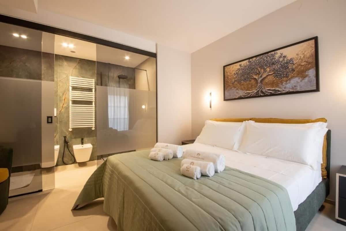 A modern hotel room with a large bed, a glass shower enclosure, and a painting on the wall.