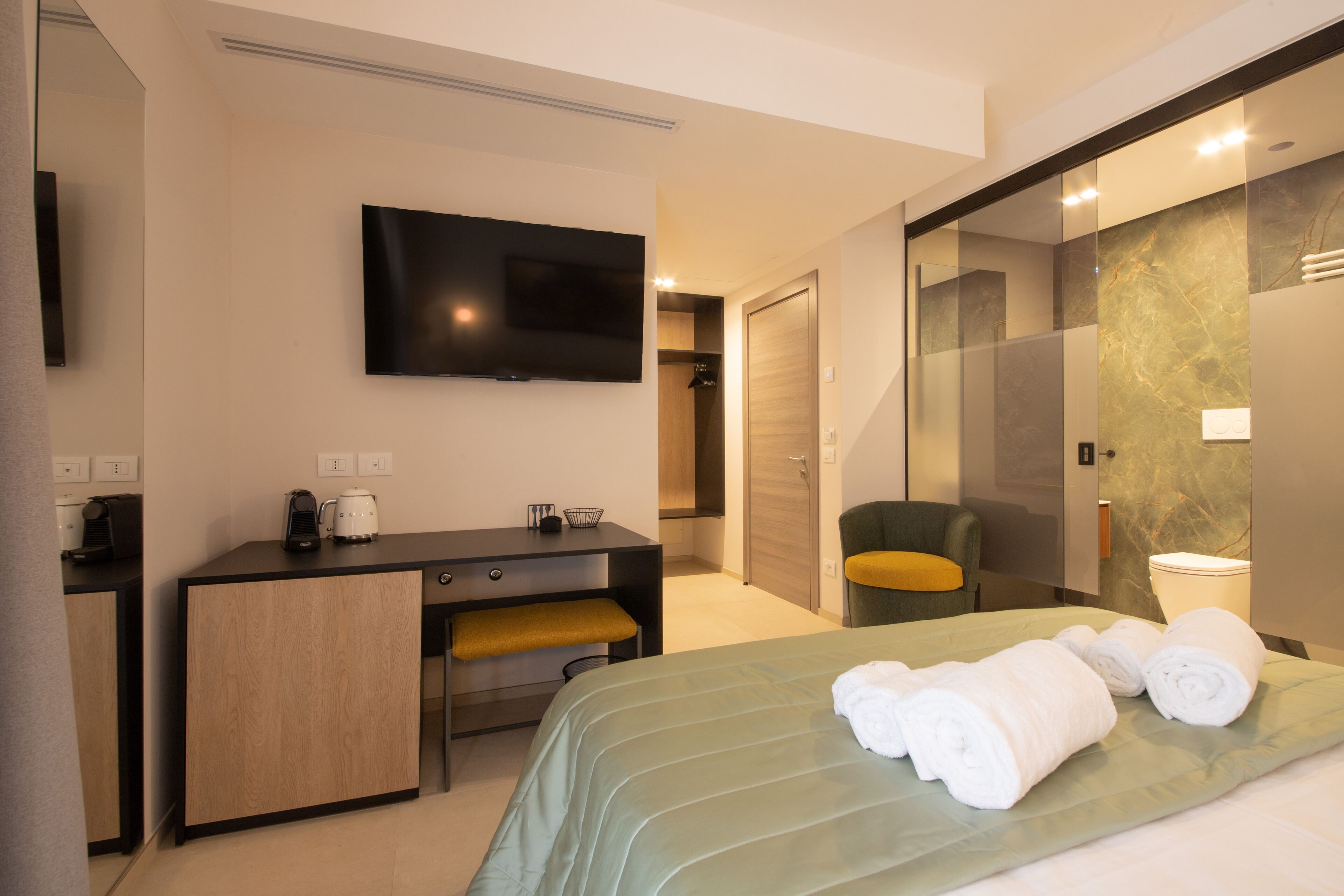 A modern hotel room with a bed, a desk, a chair, a television, and a bathroom with a glass partition.
