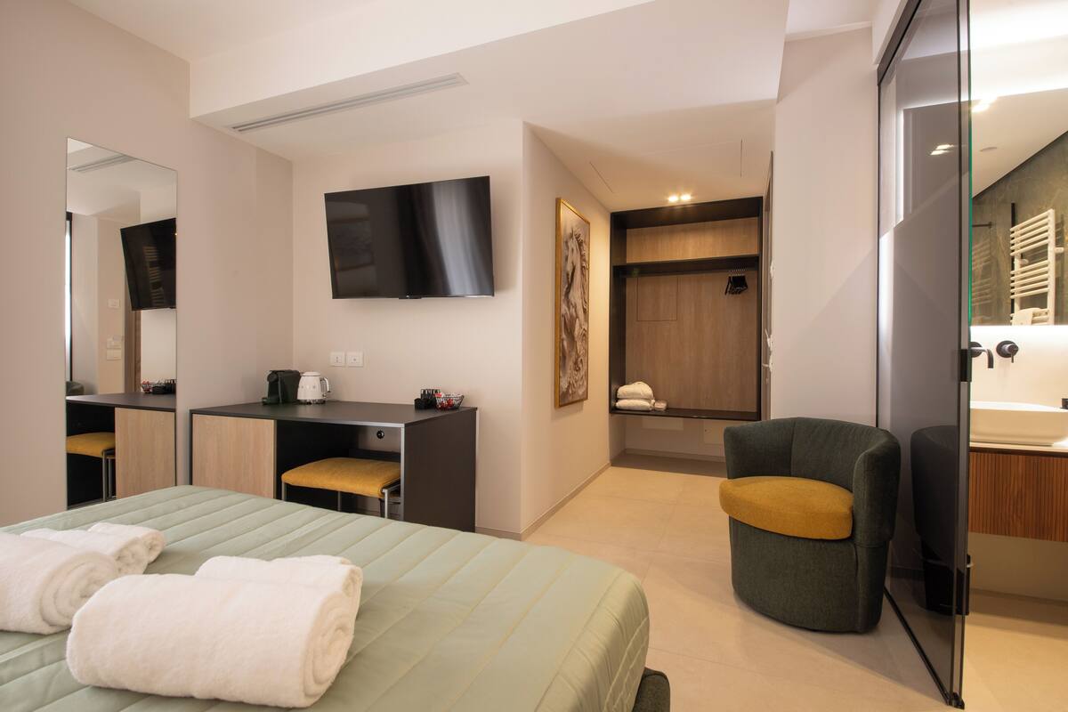 A modern hotel room with a bed, a desk, a chair, a TV, and a bathroom with a towel rack.
