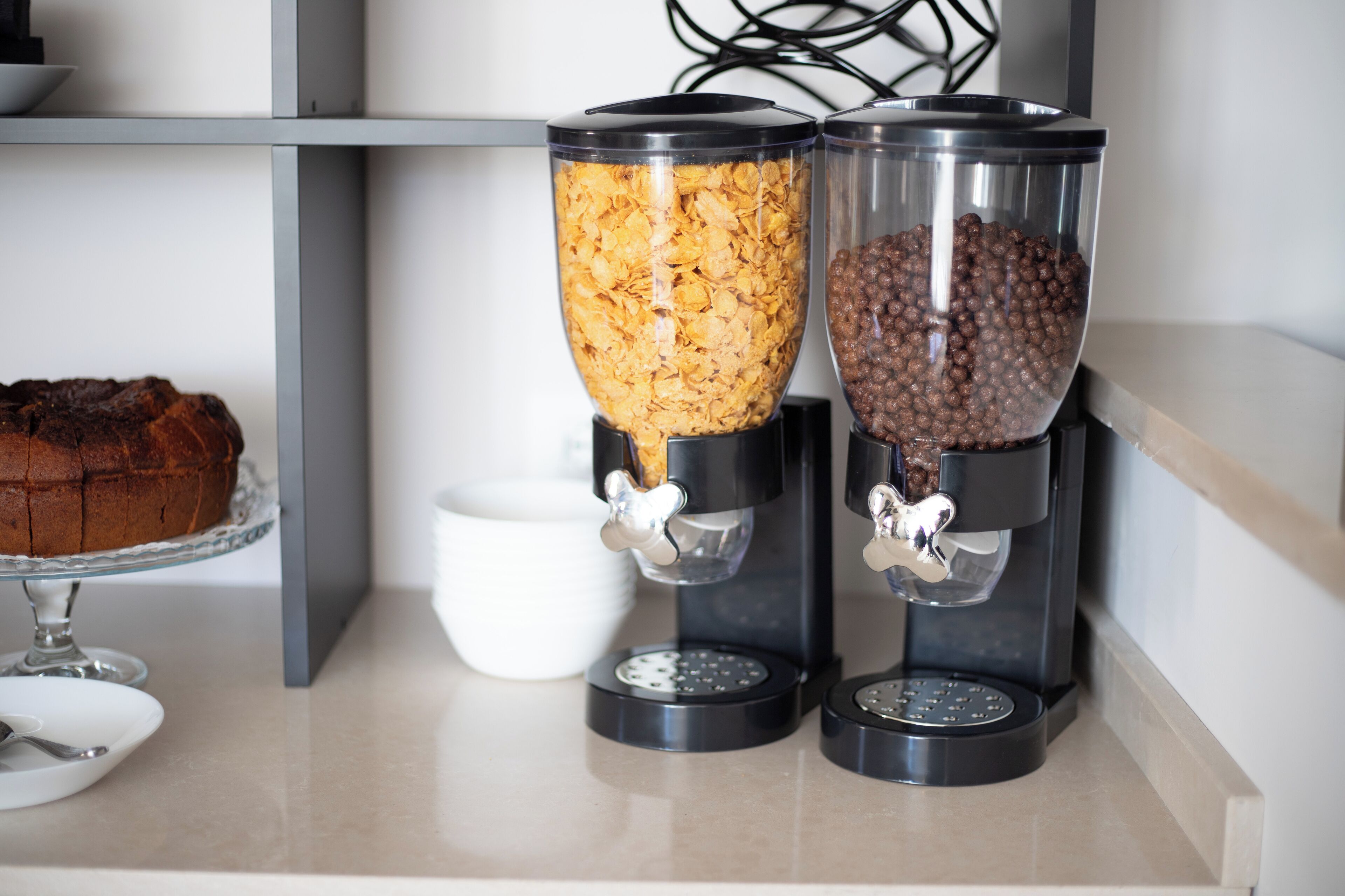 Two coffee dispensers with different beverages, a cake on a stand, and a white bowl.