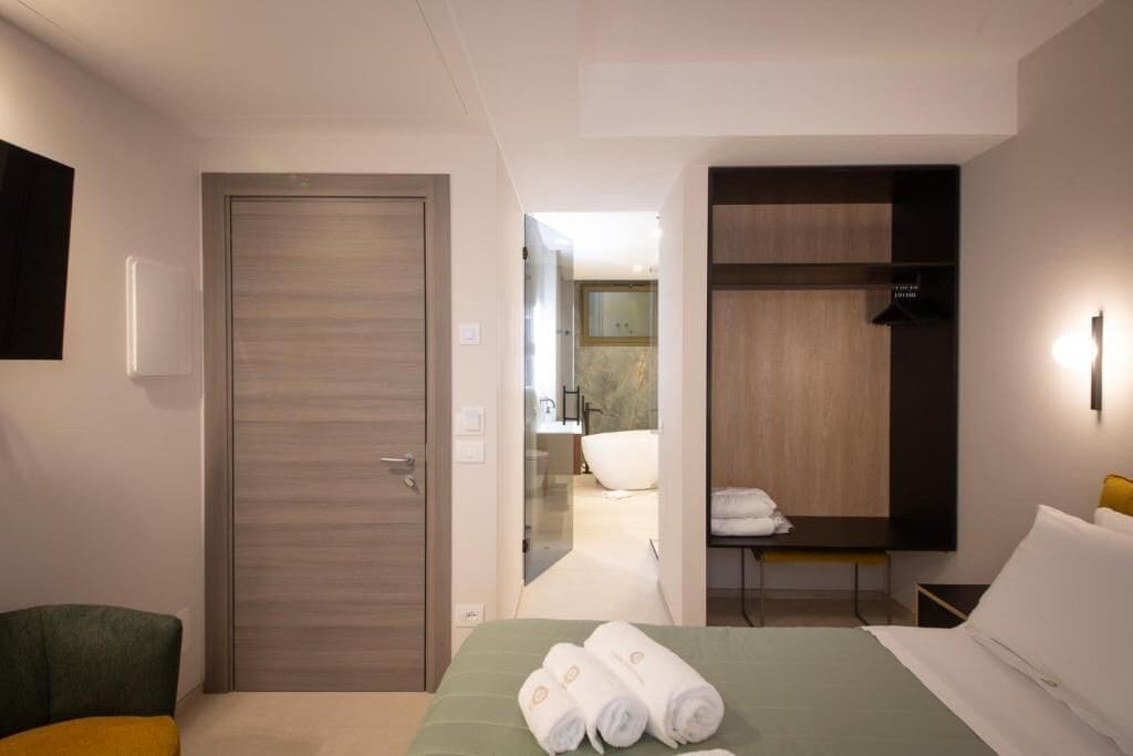Ein Hotelzimmer mit einem Bett, einem grünen Sessel, einem Holzschrank und einem durch eine offene Tür einsehbaren Badezimmer.